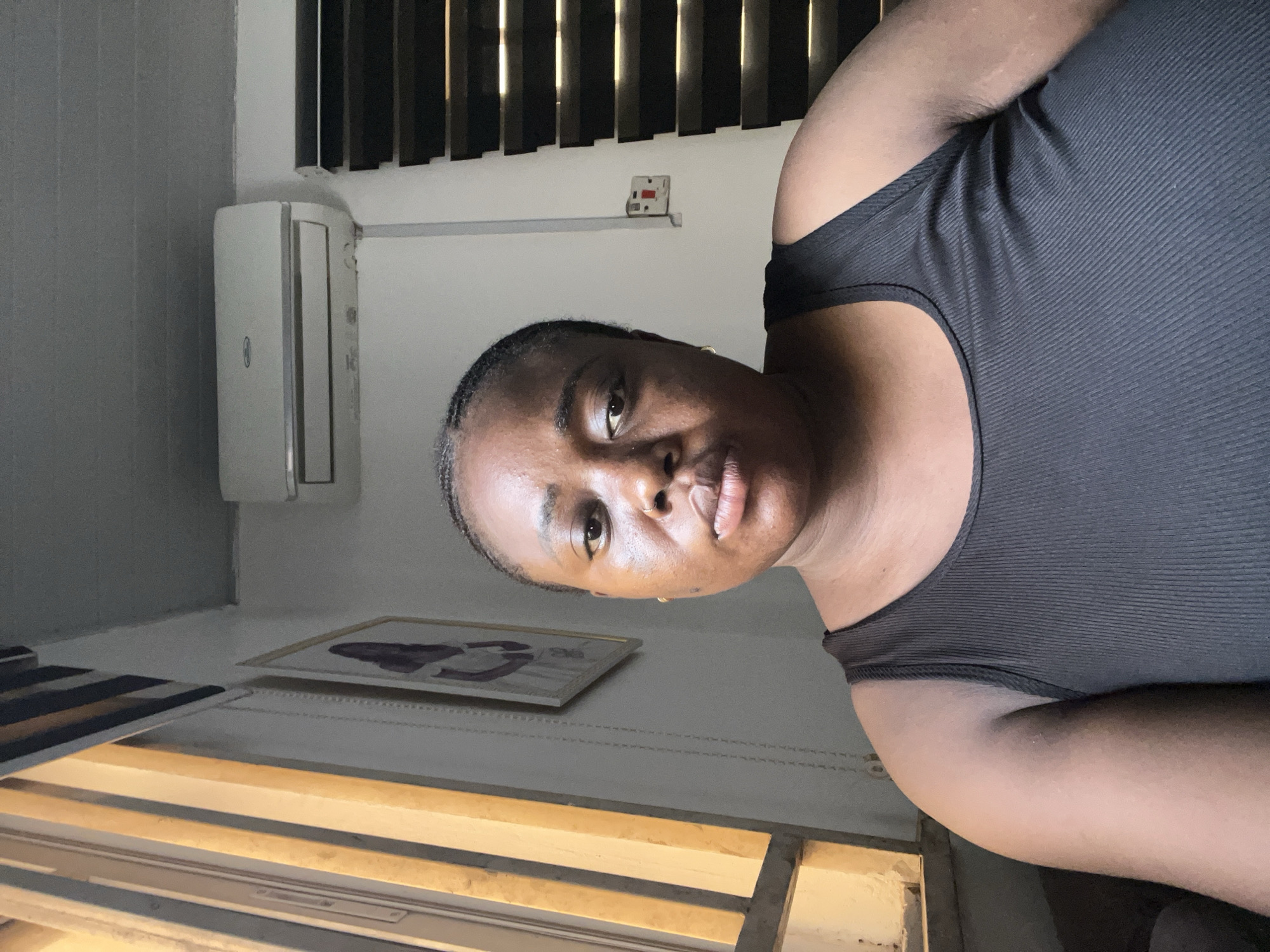 A medium shot presents a dark-skinned woman wearing a black tank top, her face directed toward the camera. The background reveals interior elements, including a wall-mounted air conditioning unit, a framed picture on the wall, and window blinds. Based on the presence of these elements, the location appears to be indoors. Information is insufficient to determine the location in the city of Abuja, Nigeria.