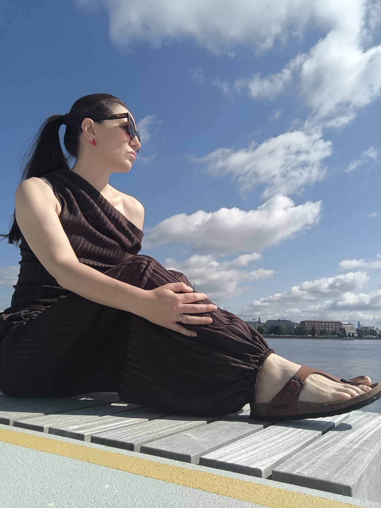 A woman with dark hair in a ponytail, wearing a brown one-shoulder top and matching pants, sunglasses, and brown sandals, sits on a wooden pier on a sunny day with a blue sky and white clouds. She is looking out at a body of water with buildings in the distance. This location appears to be the Kipsala district in Riga, Latvia, specifically near the House of the Blackheads or the Daugava River waterfront, given the architectural style of the buildings visible across the water and the presence of a pier that is typical of waterfront areas in cities like Riga. The woman's right hand rests on her left leg, and she has a silver ring on her left ring finger.