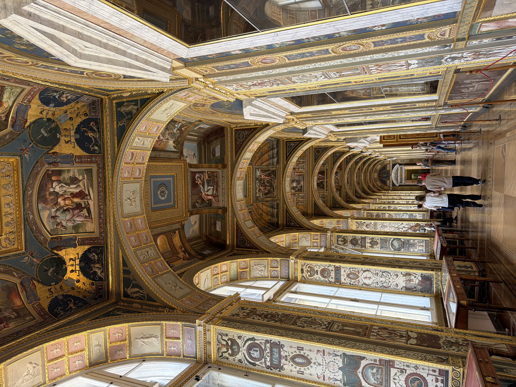The image depicts the Raphael Loggias in the Hermitage Museum, Saint Petersburg, Russia. The architectural space is characterized by a long, arched corridor adorned with elaborate frescoes on the ceiling and walls. Large windows line one side of the corridor, allowing natural light to illuminate the opulent interior. Several display tables are placed along the wall beneath the windows. Four individuals are visible in the corridor: two are walking away from the viewer towards the end of the hall, and two others are standing further down, seemingly engaged in conversation or observation. The overall impression is one of historical grandeur and artistic richness.
