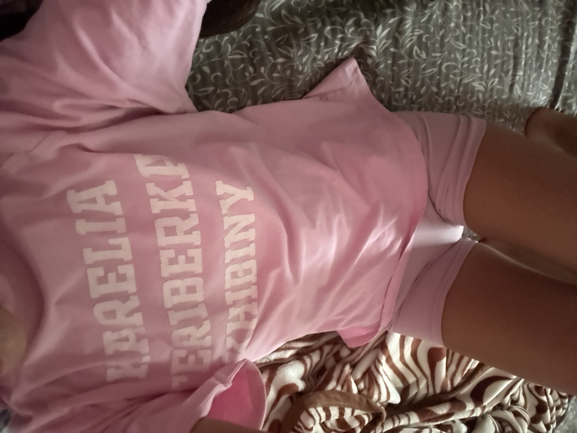 The image shows a person lying down on a bed with a pink t-shirt and pink shorts. The t-shirt has white text on it that reads "DARE TO BE FREE". The background is a patterned duvet. The image is taken from a high angle, looking down at the person. The lighting is natural, suggesting daytime. The overall mood is relaxed and casual. The location context is Basarabeasca, Moldova, but there are no specific visual cues in the image to confirm this.
