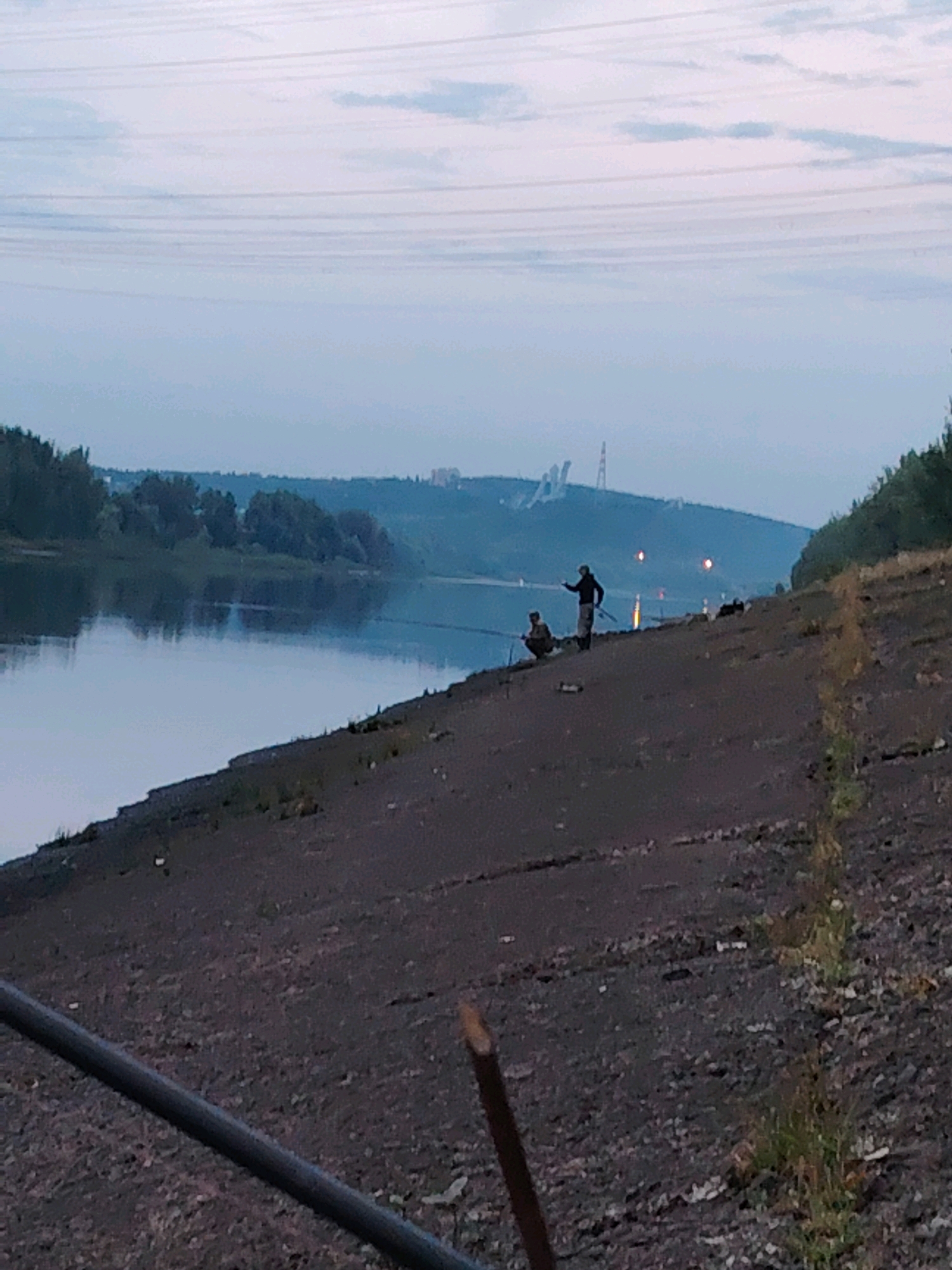 An overcast, twilight scene depicts a wide river with calm water reflecting the sky. On the near bank, a dark, sloped embankment covered in gravel and sparse vegetation leads down to the water. Two individuals are present on the embankment. One, standing, appears to be holding a fishing rod, facing away from the camera. Another individual, crouched, is also fishing. In the background, across the river, a distant, hazy skyline is visible, featuring trees, buildings, and industrial structures, including what appears to be a power line tower. The overall impression is one of quiet, early evening activity.