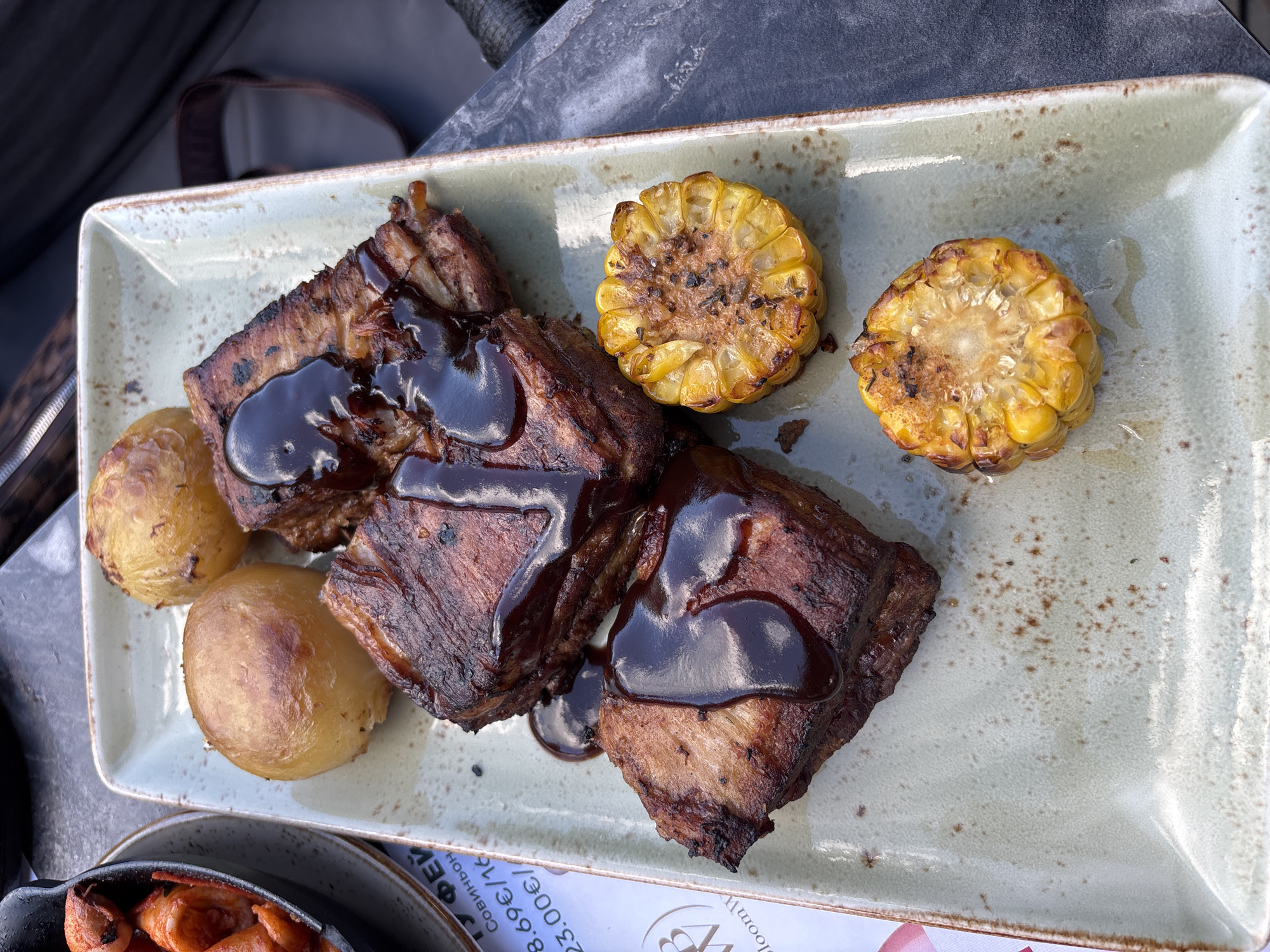A close-up, overhead shot shows a rectangular ceramic plate with a light gray speckled glaze and a brown rim. On the plate are three pieces of barbecued pork ribs, generously drizzled with a dark, glossy sauce. The ribs have a dark, caramelized exterior and appear to be tender. Accompanying the ribs are two small, golden-brown roasted potatoes and two round slices of grilled corn on the cob, with visible char marks and kernels. To the left of the plate, a portion of a black cast-iron dish is visible, containing what appears to be cooked sausage or meat. A white paper menu with Cyrillic text and a logo is also partially visible on the left side of the frame. The plate sits on a dark, textured surface, possibly a table, with a blurred background that suggests an outdoor setting. The overall scene depicts a prepared meal, likely at a restaurant. The menu text includes prices like "8.69€/16" and "23.00€/", hinting at a European location. Considering the context and style of food, this could be a scene from a restaurant in Nesebar, Bulgaria, known for its historical sites and culinary offerings.