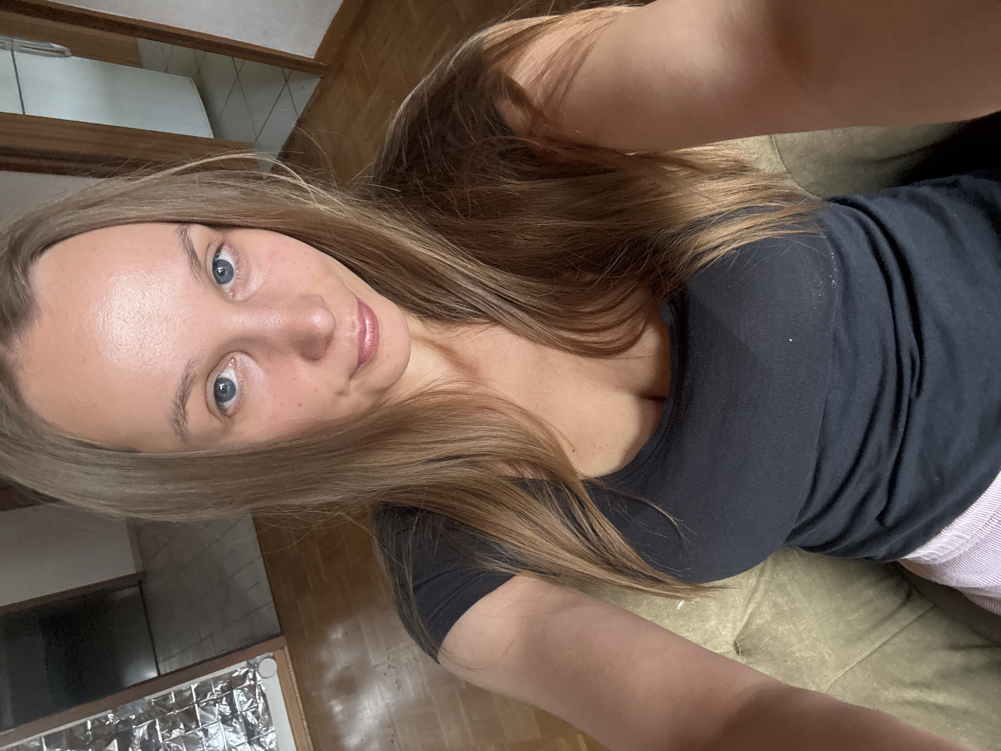 The image shows a woman sitting on a couch, taking a selfie in what appears to be a room with wooden floors and doorways visible in the background. She has long blonde hair and is wearing a black t-shirt. The setting does not provide any visual cues to indicate a specific location within the city of Überherrn, Germany.