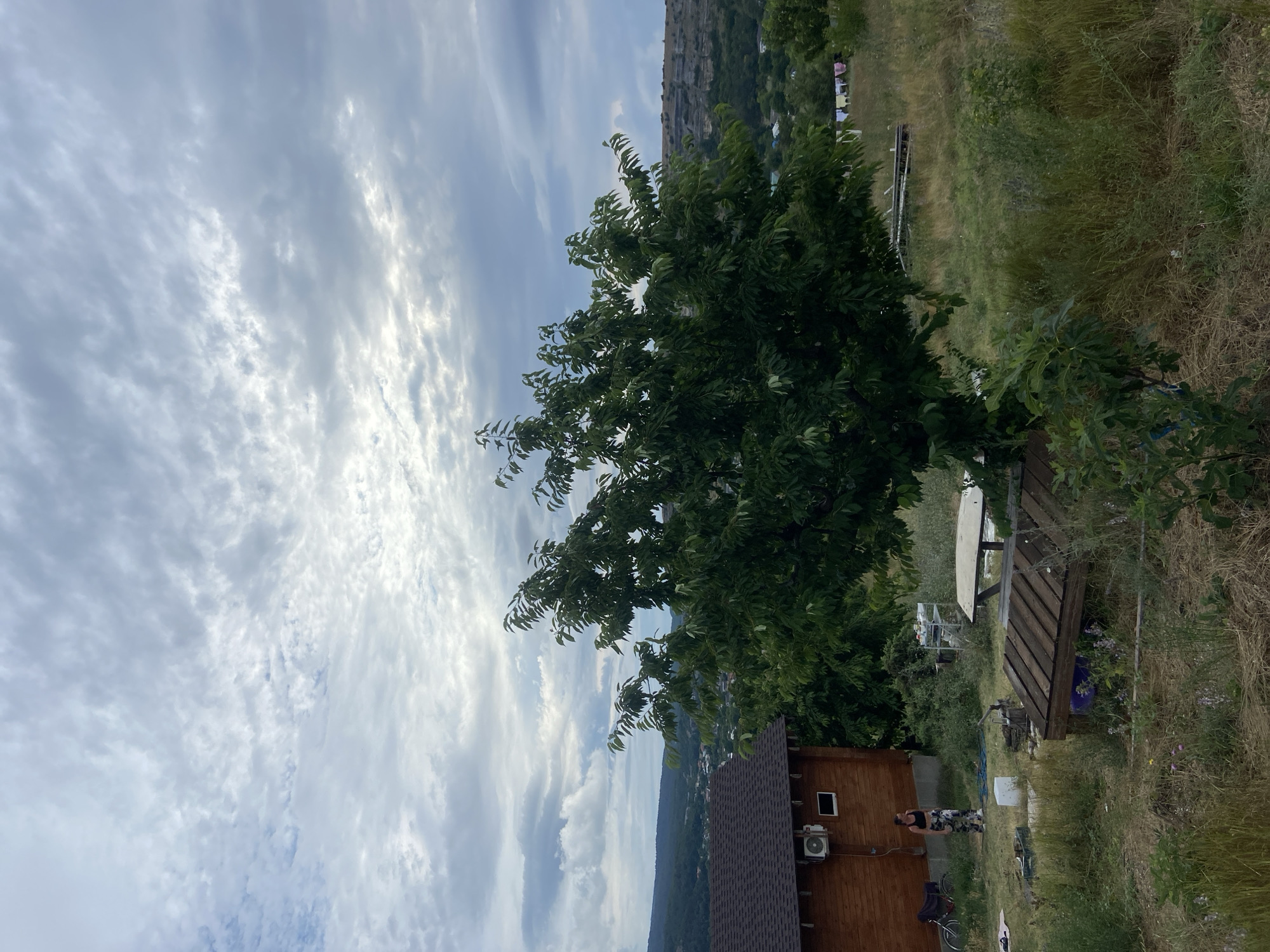 The image depicts an outdoor scene with a wooden house on the left, a large tree in the center, and a hilly landscape with a village in the background under a cloudy sky. A woman in athletic wear stands in the yard near the house, next to a bicycle. In the foreground, there is a wooden table with a bench, and other scattered objects. The environment appears to be rural, likely in or around Красномакское сельское поселение, Ukraine. The sky is filled with dramatic, textured clouds, suggesting overcast or impending weather.