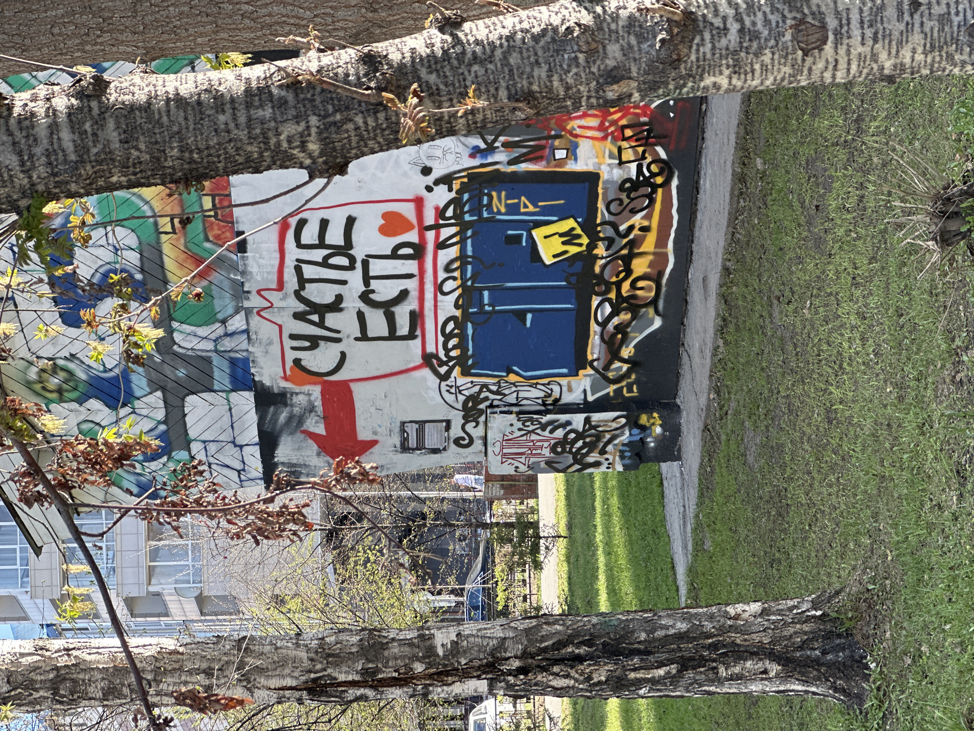 The image shows a segment of a graffitied wall next to a green patch of grass, with trees obscuring parts of the view. The wall features a variety of graffiti art, including writing in what appears to be Russian. The most prominent text reads "Счастье есть" ("Happiness exists") with a heart symbol and a red arrow pointing downwards. There's also a blue rectangular shape with "N-A-" written above, surrounded by additional scribbles and designs. The background shows buildings suggesting an urban environment. Based on the visible text and the prevalence of such street art, the location is likely within Novosibirsk, Russia.