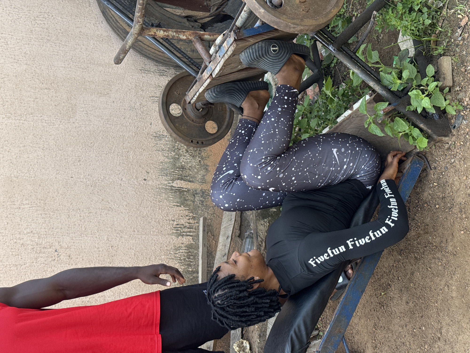 In Akwanga, Nigeria, a woman with braided hair is lying on a weight bench, performing a leg press exercise. She is wearing a black long-sleeved shirt with white lettering and patterned leggings. Her feet are on the footplate of the leg press machine, which has large weight plates attached. To her left, a man in a red shirt and black shorts stands, observing. The background shows a rough, textured wall and some outdoor vegetation. The scene appears to be an outdoor fitness area.