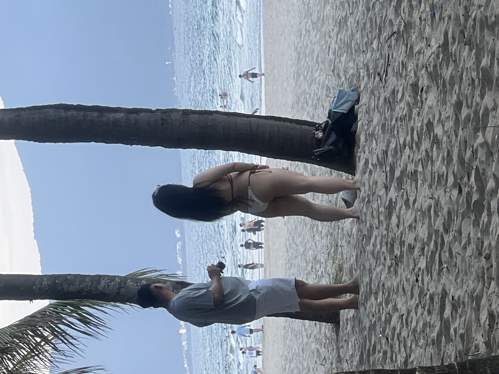 In Đà Nẵng, Vietnam, at a beach, two palm trees frame a sandy shore. The ocean is visible in the background with a hazy mountain range beyond. In the foreground, a man in a gray t-shirt and white shorts stands holding a camera. Beside him, a woman in a light-colored bikini faces away from the viewer, looking towards the sea. Other beachgoers are scattered in the water and on the sand in the mid-ground. A dark bag with a blue cloth is placed near the woman.