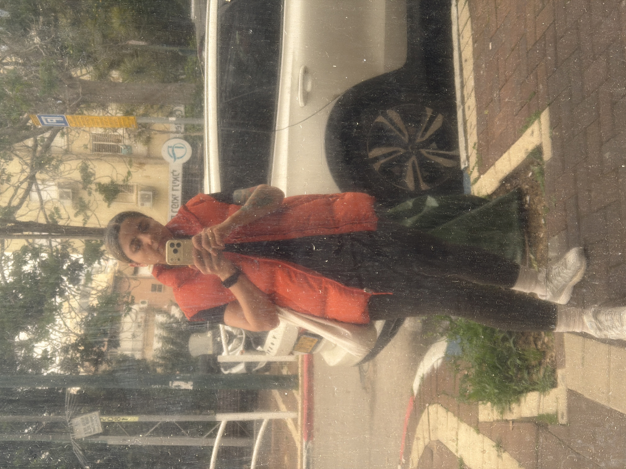 A person takes a selfie in the reflection of a window. The reflection includes a building, street signs, a white car, the person, and the sidewalk. The person appears to be wearing an orange top and black pants and is holding a cell phone. The sidewalk is adjacent to a curb with grass behind it. The street signs indicate that the location is likely Givatayim, Israel. The image appears to be taken through a dirty window.