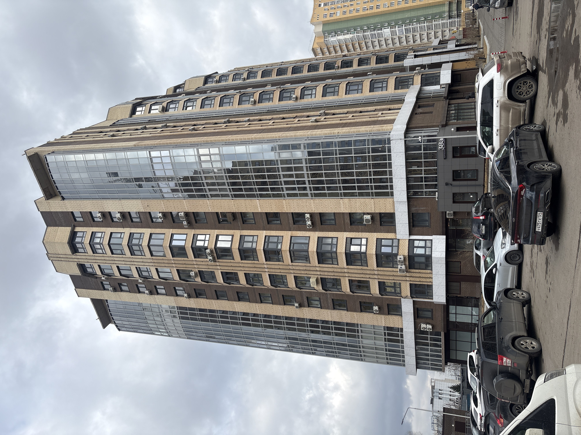 The image, captured from a street-level perspective and rotated 90 degrees clockwise, depicts a tall, modern building in an urban setting under an overcast sky. The multi-story structure showcases a distinct architectural style, combining light beige and dark brown brickwork with extensive, reflective glass sections on its lower and upper floors. The building features numerous windows, many with dark frames, and a stepped or terraced design on its upper levels, where rows of external air conditioning units are visible beneath the windows.
Along the asphalt street in the foreground, several cars are parked. These include a white SUV (possibly a Jeep Grand Cherokee), a dark sedan (like a Kia K5) with a visible partial Russian license plate "475 X 124", and a dark SUV with a rear-mounted spare tire. Some wet patches on the road suggest recent rain. Red and white striped bollards are placed on the sidewalk next to the parked vehicles.
On a dark sign near the building's entrance, vertically oriented text reads "38a" followed by the Cyrillic word "Парковая" (Parkovaya). Other buildings are faintly visible in the background against the cloudy sky. The scene captures a quiet daytime moment in what appears to be a residential or mixed-use area of Krasnoyarsk, Russia, with no people actively engaged in any events.