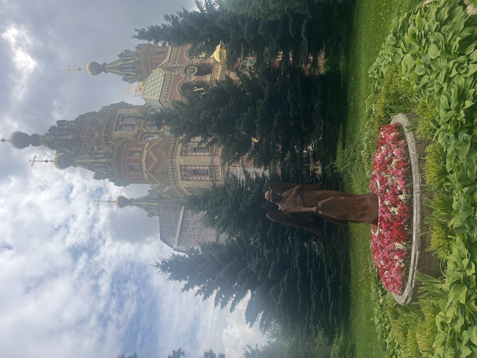 The image depicts the Peterhof Cathedral, a prominent landmark in Peterhof, Russia, situated against a cloudy sky. In the foreground, a flowerbed bursting with red, pink, and white blossoms surrounds the base of a bronze statue of an angel. The angel, rendered in a stylized, angular form, stands with outstretched wings, holding an object in its left hand. Lush green grass and large-leafed plants frame the flowerbed, creating a vibrant natural setting. Behind the statue, tall, dark green fir trees partially obscure the view of the cathedral, their dense foliage creating a striking contrast with the lighter tones of the sky and the cathedral's architecture. The cathedral itself, with its ornate red brick facade, multiple golden domes topped with crosses, and intricate detailing, stands majestically in the background, hinting at its historical and religious significance. A clock face is visible on one of the cathedral's towers. The overall atmosphere is one of serene grandeur, with the natural beauty of the garden complementing the architectural splendor of the cathedral.