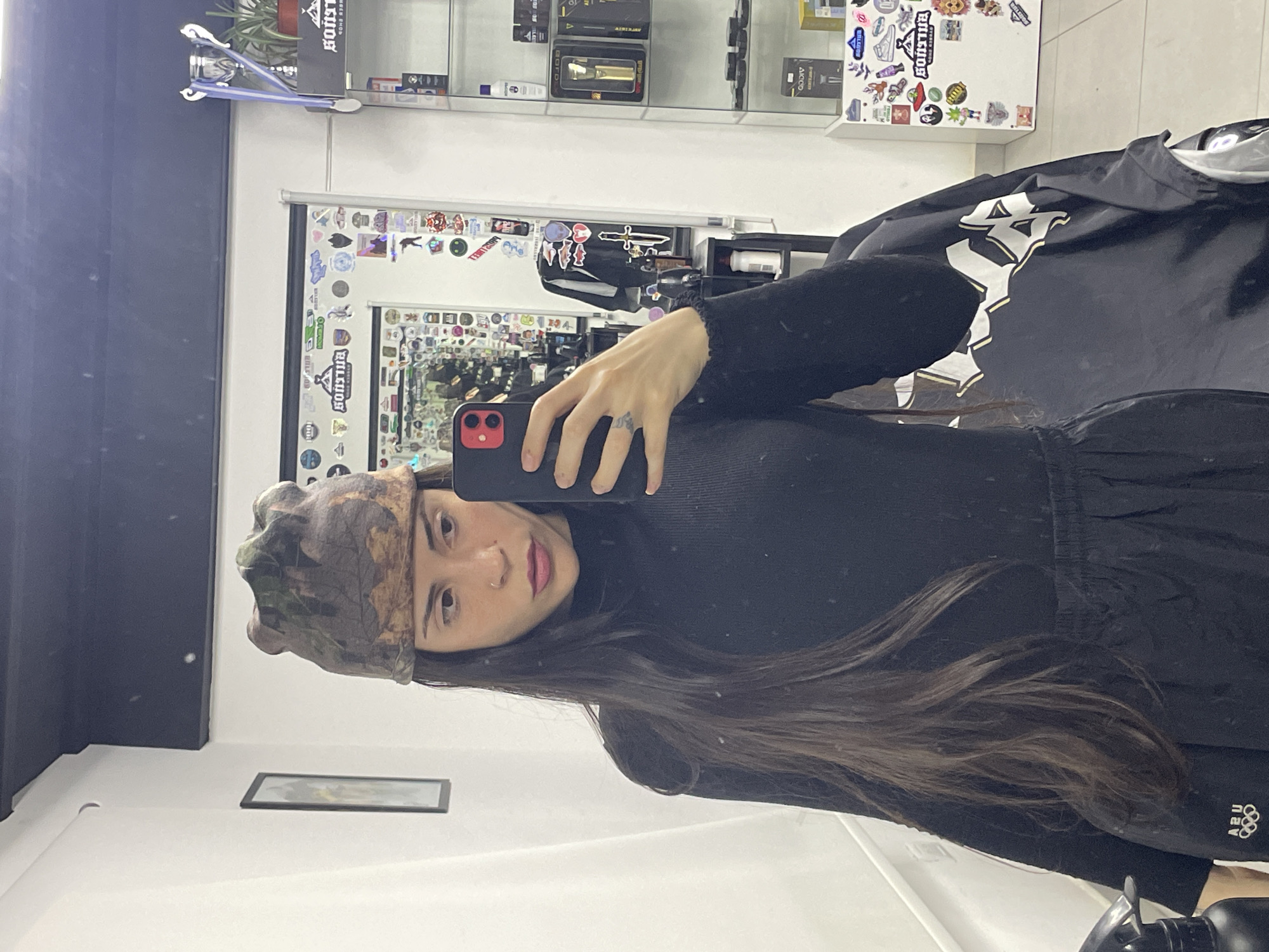 A woman wearing a camouflage beanie and black clothing is taking a selfie in a barbershop. She is holding a red phone. A black barber's cape is partially visible. The barbershop features a mirror decorated with various stickers, and shelves displaying products. The location is likely Ushuaia, Argentina, based on the interior design elements commonly found in similar establishments in that city.