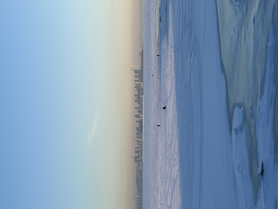 The image presents a wide-angle, eye-level view of a frozen winter landscape looking towards the city of Saint Petersburg, Russia. The scene is situated on the Gulf of Finland or Neva Bay, with the urban skyline visible on the distant horizon.
The sky, occupying the upper third of the frame, is clear, transitioning from a light blue overhead to a pale yellow-orange haze at the horizon. A faint, wispy cloud is discernible in the upper-center sky.
The middle ground is dominated by a vast, snow-covered expanse of ice, exhibiting numerous faint tracks and patterns. Four individuals are present on this frozen surface: one person stands in the mid-left, another is kneeling next to a dark, rectangular, portable shelter (suggesting ice fishing) in the mid-center, a third person stands further back to the right, and a fourth person stands far right, closer to a darker area of water or thinner ice.
The distant horizon showcases the Saint Petersburg skyline, characterized by multiple high-rise buildings, including the prominent, slender Lakhta Center tower visible on the far right. Industrial structures, cranes, and a large, dark vessel (possibly a ship or barge) are also present along the distant shoreline, partially obscured by atmospheric haze.
The immediate foreground features a blend of snow-covered ice and distinct patches of darker, reflective water or thinly frozen ice, displaying irregular cracks and formations. A small, jagged piece of ice floats in a patch of water in the lower-left foreground. The overall lighting suggests conditions typical of early morning or late afternoon.