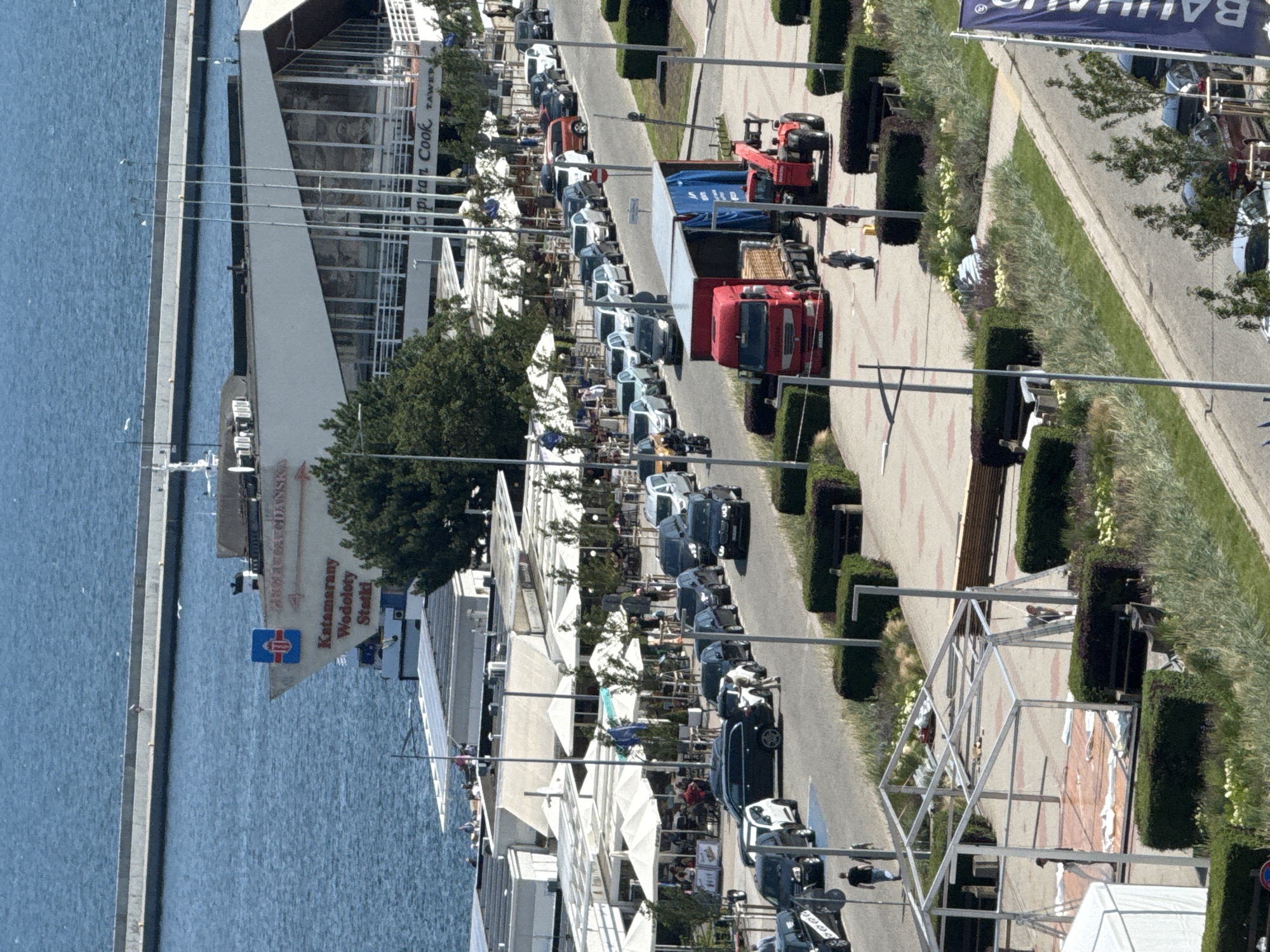 This aerial view captures a bustling scene along the waterfront in Gdynia, Poland, specifically near the Gdynia Główna railway station and the Gdynia Aquarium. In the background, the blue waters of the Baltic Sea meet a long, concrete breakwater. Prominently displayed on a building is the text "Gdynia Główna" and "Kataramany Wodoloty Statki," indicating a ferry terminal. Several smaller buildings with white awnings, likely cafes or restaurants, line the waterfront.
In the mid-ground, a street is filled with parked cars and a red truck with an attached trailer. A red forklift is positioned beside the trailer, suggesting activity related to loading or unloading. People are visible walking along the sidewalk and near the vehicles. Further down the street, a line of cars is parked.
The foreground shows a construction area with a partially assembled metal structure, possibly a tent or canopy, with workers present. Various green bushes and landscaping elements are interspersed throughout the scene, indicating a well-maintained public space. A large blue banner with white text, partially visible on the right edge of the frame, reads "BAUHAUS," suggesting a nearby retail establishment. The overall impression is one of a vibrant urban area experiencing a high level of activity, likely during a warm day.