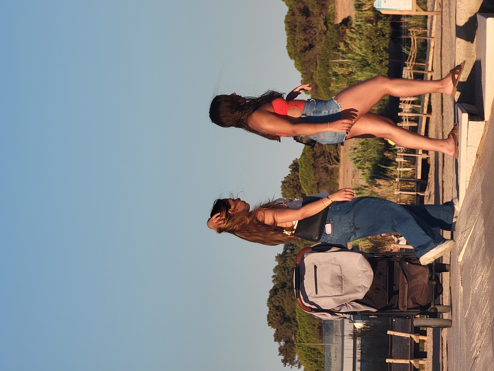 Two young women walk on a wooden pathway in Quarteira, Portugal. The woman on the left, with long brown hair, wears a grey tank top and blue jeans, carrying a black purse. She has her hand in her hair. Beside her is a grey stroller with brown accents. The woman on the right, with long dark hair, wears a red one-piece swimsuit and denim shorts. She is walking on a raised concrete structure with a wooden railing. In the background, a line of trees and vegetation is visible under a clear blue sky.