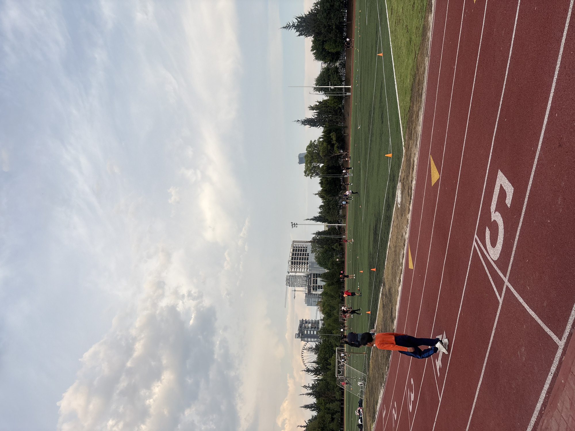This image captures an outdoor sports complex, featuring a red running track that encircles a green athletic field. In the foreground, a person wearing an orange long-sleeved shirt, dark pants, and white shoes stands on lane 5 of the running track. White lines delineate the lanes, and yellow triangular markers are visible on the track. The number "5" is clearly painted in white on the track surface.
Beyond the track lies a lush green field, likely used for football or soccer, dotted with several orange cones, suggesting training or practice. Numerous individuals are scattered across this field, some appearing to be engaged in sports or exercise activities. The field is bordered by a line of trees. In the background, an urban landscape unfolds, with multiple buildings, some clearly under construction, and a prominent Ferris wheel adding to the skyline.
The sky is partly cloudy, displaying a mix of light and darker clouds, indicating a late afternoon or early evening setting with soft, natural lighting. The scene conveys an active yet calm atmosphere at a sports facility. This location is consistent with an urban or suburban area, specifically identified as Puebla, Mexico.