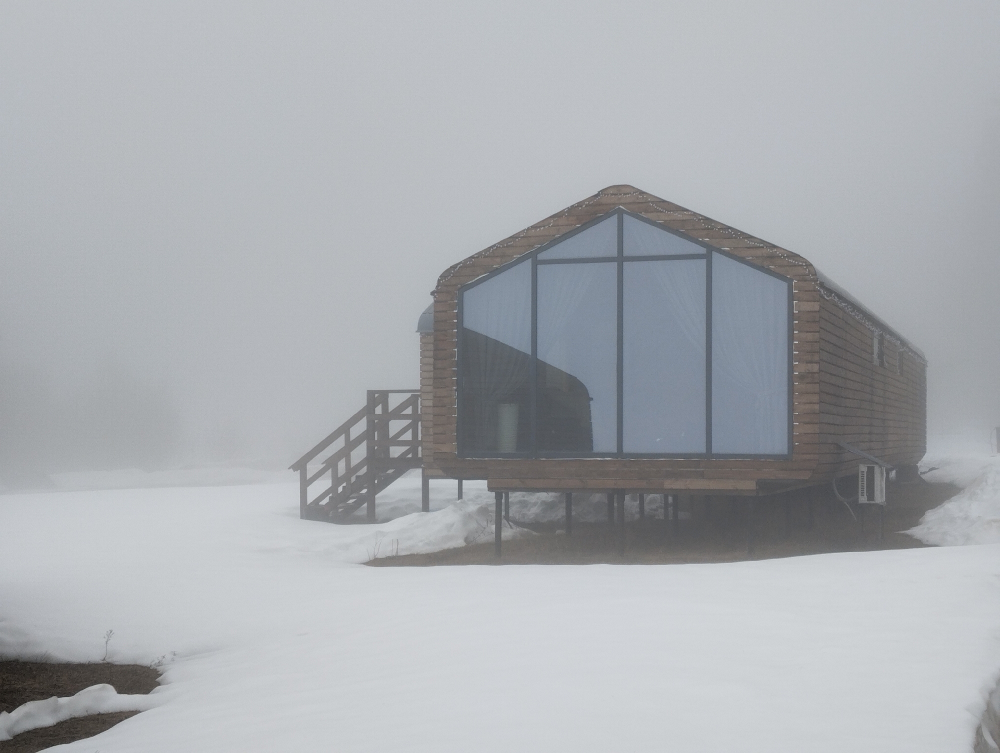 A modern cabin sits on a snow-covered landscape under a heavy fog. The cabin has a distinctive A-frame design with a large glass window facade. The structure is raised on posts, and a wooden staircase leads to the entrance. An air conditioning unit is visible on the side of the cabin. Snowdrifts surround the base of the structure. Given the presence of modern architecture and snowy conditions, it is plausible this scene takes place in Votkinsk, Russia. There are no people visible in the image.