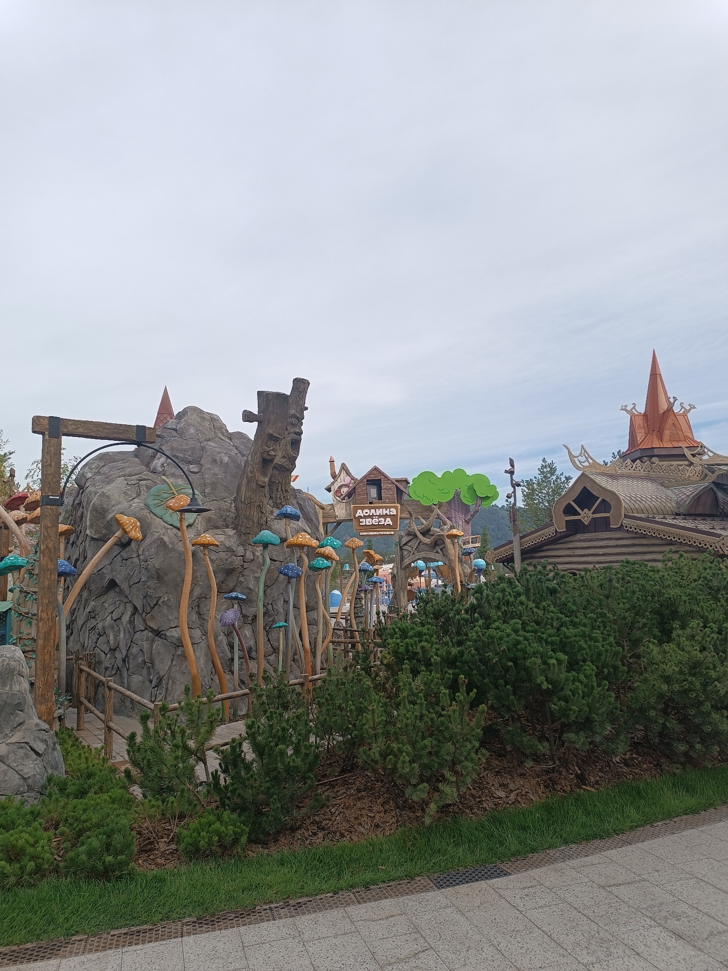 A medium-wide shot shows an outdoor scene with a whimsical theme, likely an amusement park or a themed attraction. In the foreground, a paved walkway with a grate runs horizontally across the bottom of the frame. Behind this, a strip of green grass is bordered by dense evergreen bushes.
The mid-ground is dominated by a large, grey, rocky structure that resembles a cliff face, adorned with numerous colorful, oversized mushroom sculptures on wooden stems of varying heights. Some mushrooms are a vibrant blue, others orange, teal, and purple. A wooden walkway and railings are visible, suggesting access to or around this area. A large, gnarled tree stump with a carved face is integrated into the rock formation. To the right of the rock, a building with a steeply pitched, copper-colored roof and intricate wooden trim is visible. This building features carved ornamentation and a spire-like structure on its peak. Another wooden building with a pitched roof and a decorative gable is situated further to the left. A sign with Cyrillic text, "ДОЛИНА ЗВЁЗДА" (Valley of Stars), is mounted on a wooden archway. Behind these structures, a stylized green tree shape is prominent. The sky above is overcast, with diffused light. No people are visible in the image.
