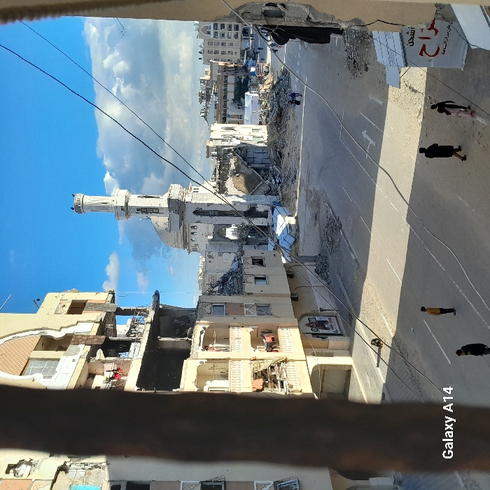 This image depicts a street scene in Khan Yunis, Palestinian Territory, characterized by extensive structural damage under a partly cloudy blue sky.
The foreground is partially framed by fragments of two damaged buildings; a brown structural element occupies the left, and a wall section is on the right. In the midground, a wide, asphalt road features two white directional arrow markings. To the left of the road, a multi-story building exhibits severe damage, with exposed interiors across several floors, shattered windows, and debris; clothing items are visible hanging from balconies. Opposite, on the right, a partially visible building also shows structural damage, with a sign containing Arabic text visible at its base.
Dominating the midground is a large, white mosque with a distinctive minaret and dome, both showing visible damage and structural compromise to their facades. The area surrounding the mosque and extending across the mid-distance is covered in substantial piles of rubble and debris from collapsed structures. Further buildings, some appearing damaged, line the distant horizon.
Six individuals are present on the street. Four figures are in the foreground-midground: one person in a yellow shirt stands centrally, facing right; one person in dark clothing walks away from the viewer, accompanied by a smaller figure in light attire; and another person in dark clothing walks towards the left. Two additional figures are discernible walking further down the street near the debris piles. Multiple utility wires traverse the scene overhead. The watermark "Galaxy A14" is visible in the bottom-left corner.