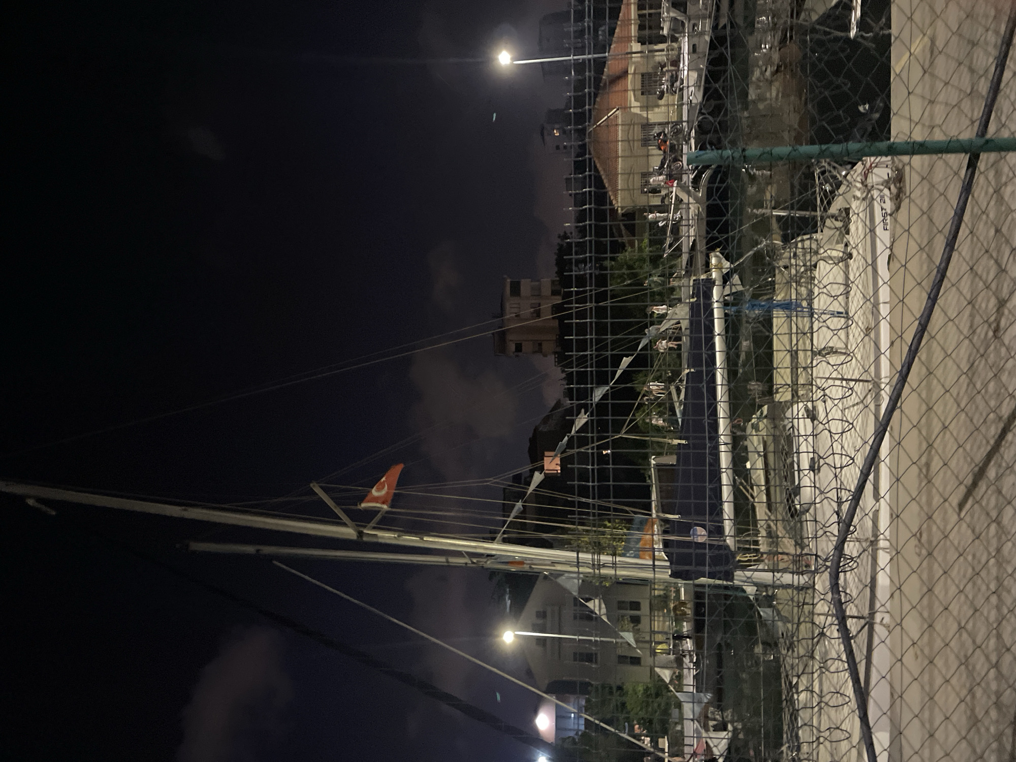 A nighttime, medium-shot image captures a harbor in Kadıköy, Turkey, with several sailboats docked. A chain-link fence runs across the foreground, partially obscuring the view. The sky is dark, with some clouds visible. The masts of sailboats are prominent, with rigging and sails visible. One sailboat features a Turkish flag. In the background, buildings are illuminated, and streetlights cast a glow. A few people are visible on the docks and near the boats. The scene appears to be a quiet evening at a marina.