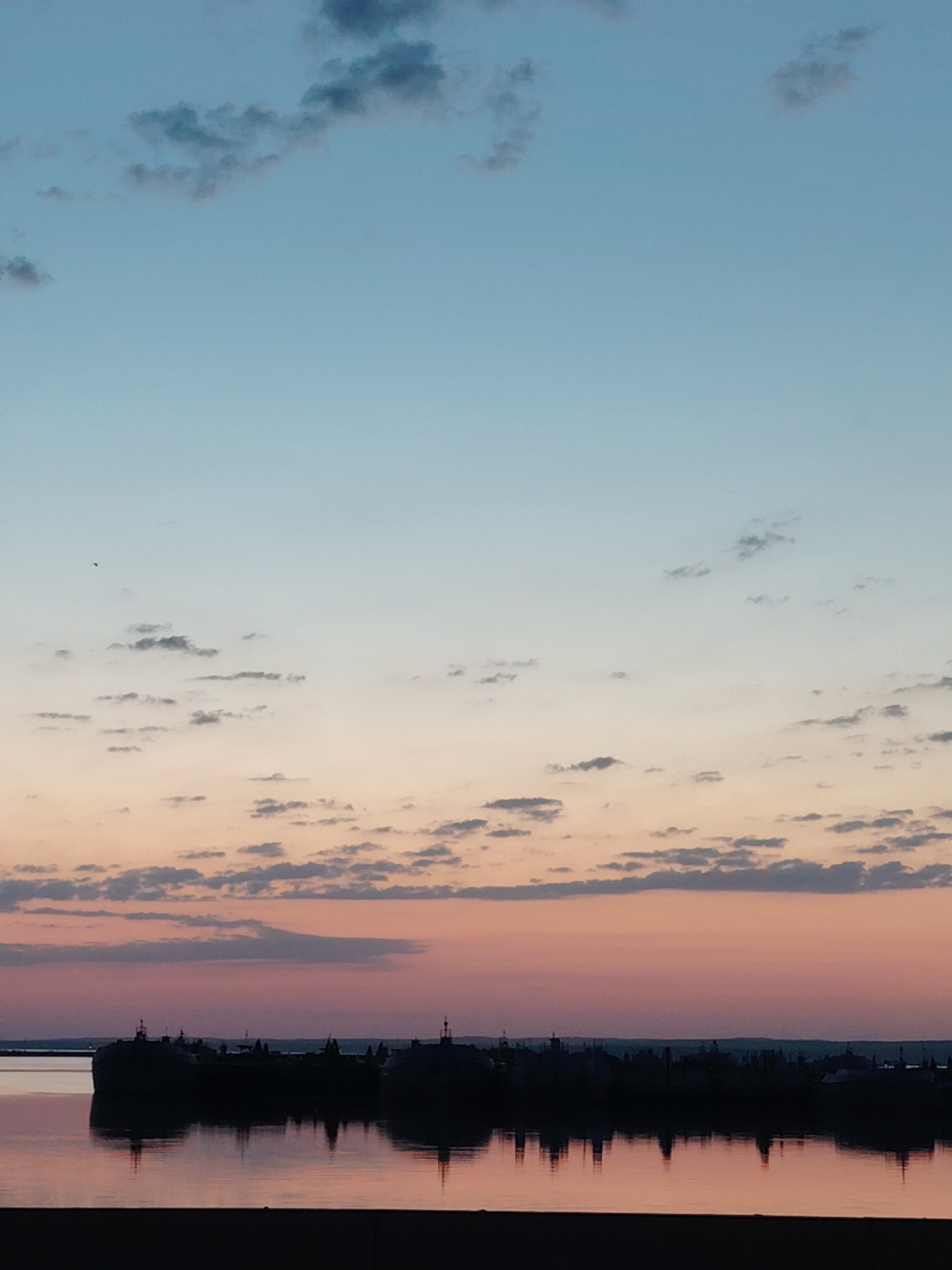 The image captures a twilight scene in Chaikovsky, Russia, showcasing a serene waterscape. Silhouetted structures, likely buildings or boats, are reflected in the calm water, creating symmetrical patterns. The sky transitions through shades of blue, peach, and pink, dotted with scattered clouds. The horizon features a darker line, possibly indicating land or distant objects.