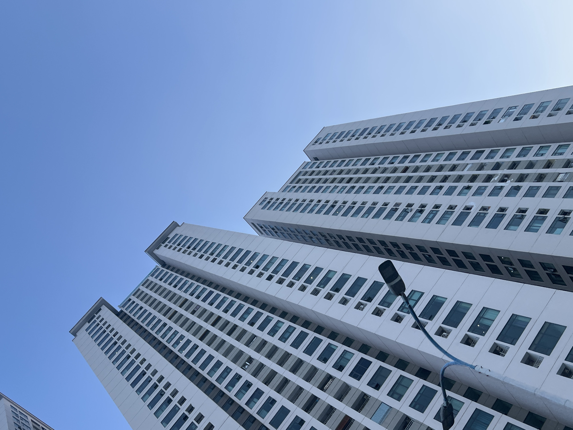 The image depicts a low-angle view of multiple modern, white high-rise buildings with repetitive window patterns against a clear, blue sky. A lamppost with curved arms is visible in the foreground, partially obscuring the building on the right. The location is Bac Nha Trang Ward, Vietnam.