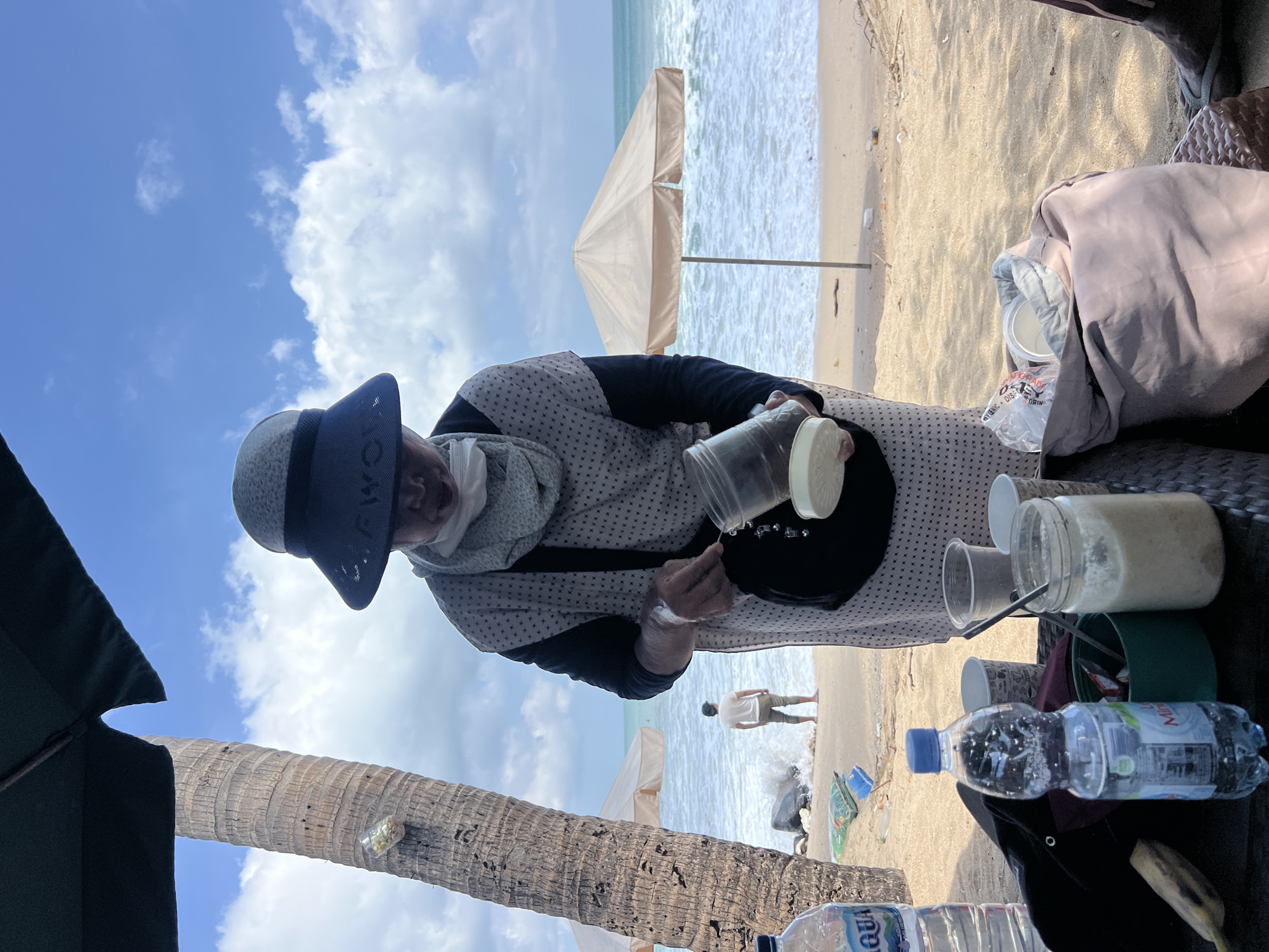 A person wearing a wide-brimmed hat and a face mask is standing on a sandy beach in Kuta, Indonesia, holding a glass jar. The person is wearing a patterned dress and a scarf. In front of the person are several bottles of water, cups, a green container, and a jar with a light-colored substance. In the background, the ocean with waves is visible, along with a person standing near the water, and two beach umbrellas. A palm tree is on the left side of the image. The sky is blue with scattered clouds.