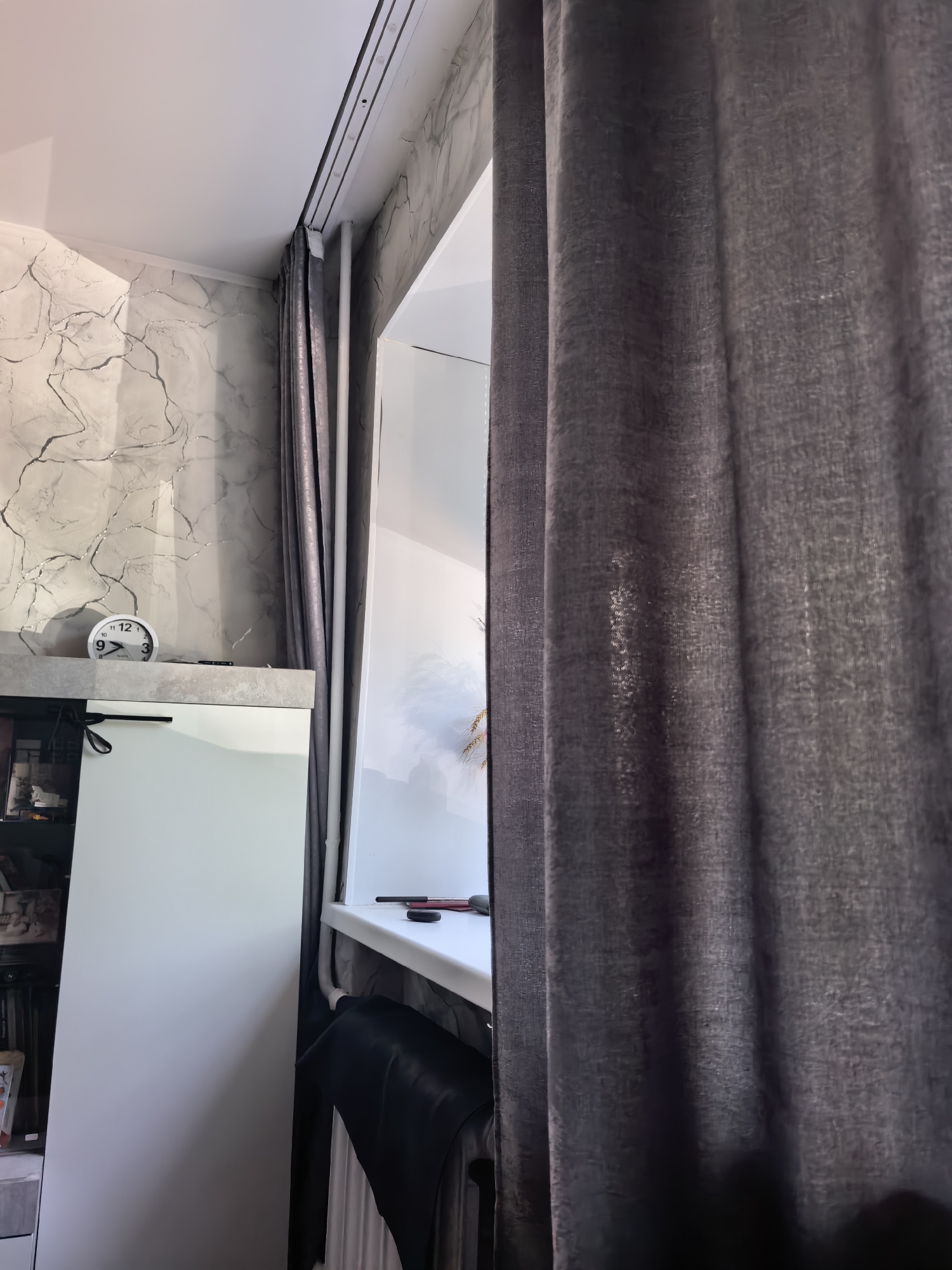 The image shows an interior view of a room, potentially located in Sterlitamak, Russia, given the prompt's instructions. The visible elements include:
* **Curtains:** Dark gray or brown curtains dominate the right side of the image.
* **Window:** A window is partially visible behind the curtains.
* **Window Sill:** A white window sill is present with objects (possibly pens/markers) and a small black item on it.
* **Cabinet:** A white cabinet is on the left side of the image, with an analog clock on top.
* **Wall:** The wall has a gray and white marble-like design.
* **Chair:** A black chair is partially visible near the window.
* **Other Objects:** Part of a shelf with items are on the left of the image.
The scene suggests a room with typical furnishings and decor. No people are visible. The lighting seems subdued, likely from natural light coming through the window.