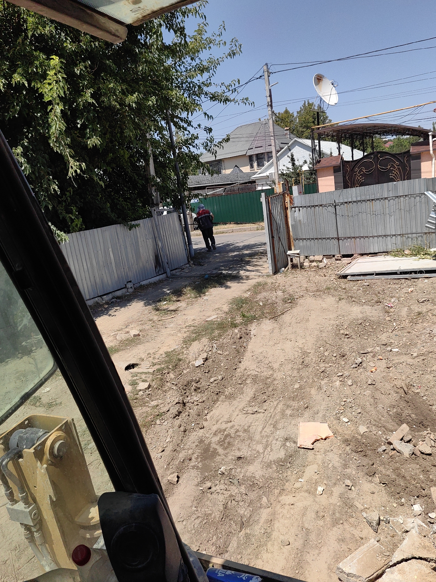This image depicts an outdoor scene, likely in a construction or renovation area, within the city of Shymkent, Kazakhstan. In the foreground, the interior of a yellow heavy machinery vehicle is visible, with a black frame obscuring a portion of the view. The ground is a mixture of dirt and debris, including scattered bricks and a piece of orange foam insulation. A person wearing a red and black vest and a blue cap is walking away from the viewer down a dirt path towards a metal gate. Behind the gate, there are houses with tiled roofs and a satellite dish mounted on a pole, characteristic of residential areas. A tall green fence runs along one side of the path, and a corrugated metal fence is also present. The sky is clear and blue.