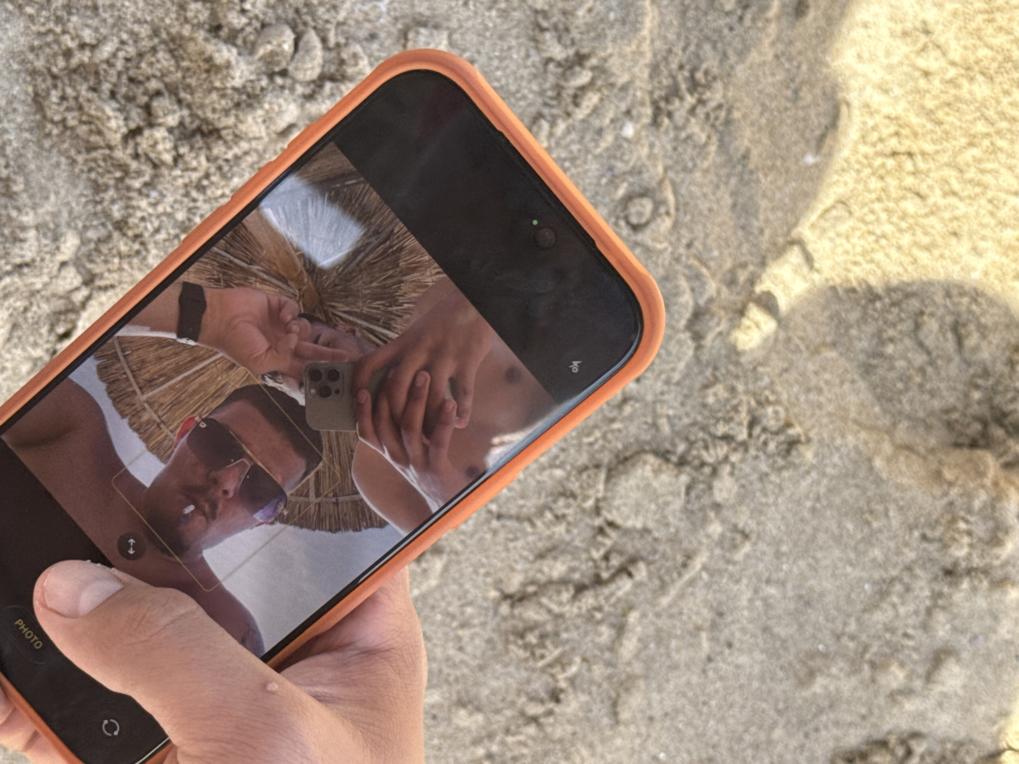A downward-angled shot captures a light-skinned hand holding a smartphone, encased in an orange protective cover. The phone's screen displays a clear reflection of a shirtless individual (likely male) wearing sunglasses, holding a second smartphone with a multi-lens camera array, appearing to take a selfie. A dark-banded watch is visible on the reflected individual's left wrist. In the background of the reflection is a straw beach umbrella. A small water droplet rests on the screen near the thumb of the hand holding the phone. The thumb and index finger of the hand are visible, with a small blemish present on the back of the index finger. The entire scene is set against a textured, light-brown sandy surface, characteristic of a beach, with direct sunlight casting distinct shadows in the lower right portion of the frame. This beach setting is consistent with coastal areas in Varna, Bulgaria.
