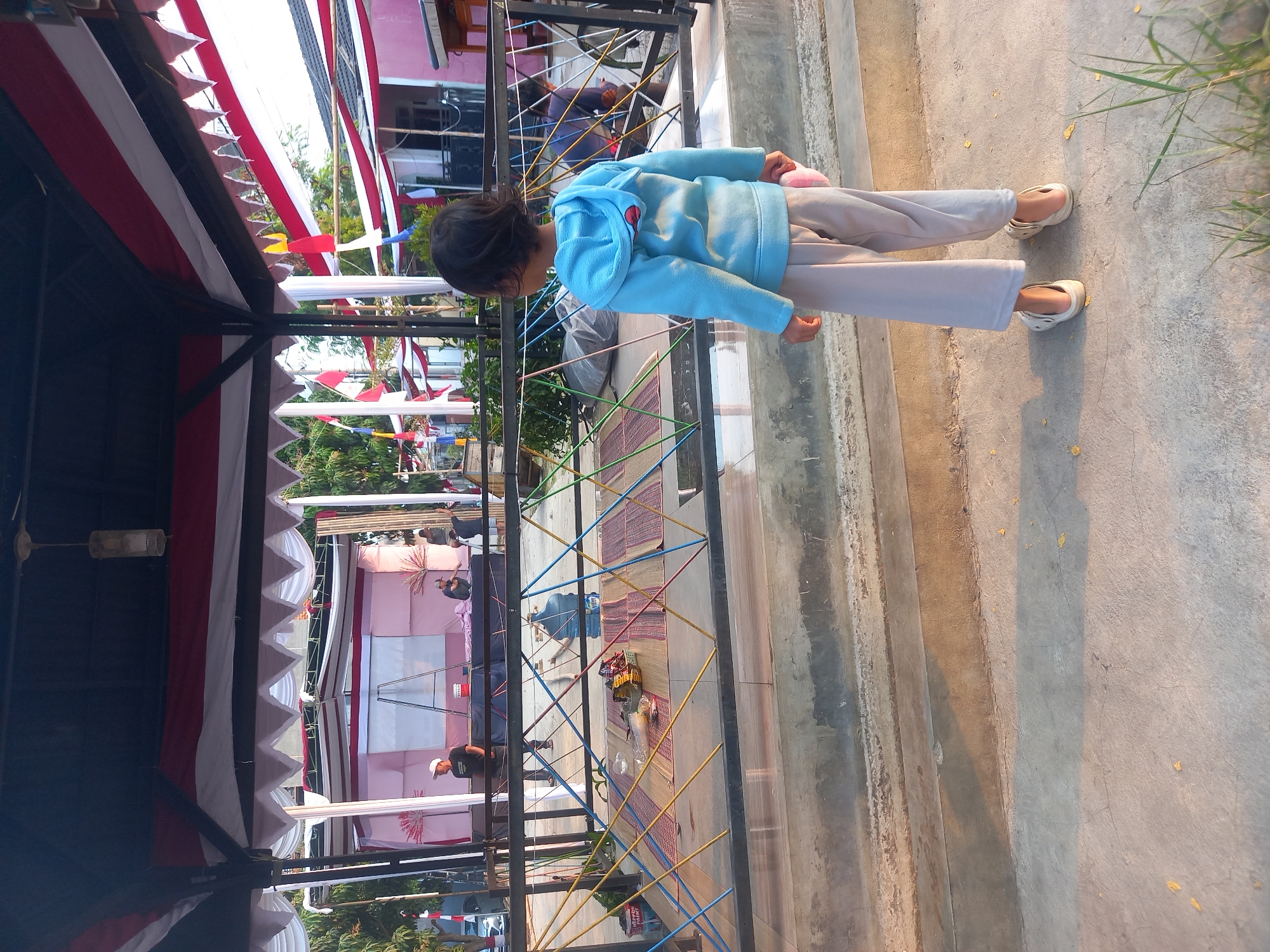 The image shows a young child, seen from behind, wearing a blue hoodie and light-colored pants. The child is standing on a concrete surface facing a decorated stage area. The stage appears to be set up for an event, with red and white draped fabric overhead and along the sides, reminiscent of Indonesian national colors. There are people visible on and around the stage, possibly setting up or preparing for an event. In the foreground, a black metal railing separates the child's viewing area from the stage. Behind the railing, mats are laid out on the ground, and a large water dispenser is present. The overall environment suggests an outdoor gathering or celebration in Cankuang, Indonesia.