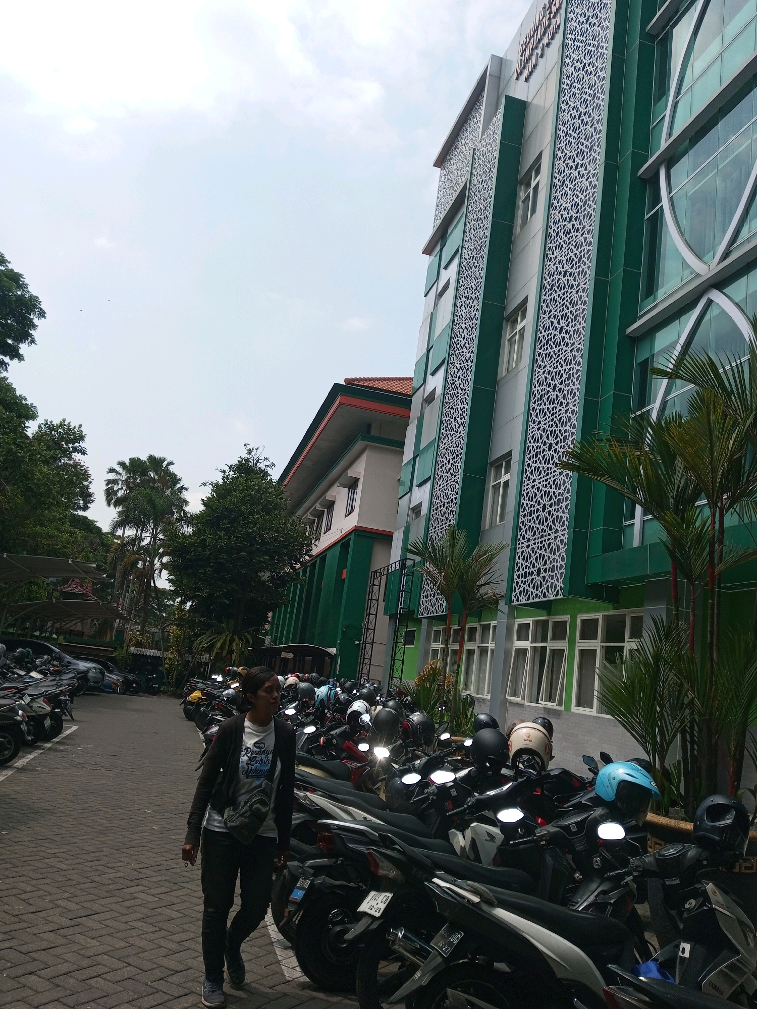 A young woman with dark hair tied back walks past a row of parked motorcycles in what appears to be a parking lot. She wears a dark jacket over a white t-shirt and dark pants, with a grey bag slung across her body. The motorcycles are parked in front of a modern building with green accents and decorative white latticework on its facade. Palm trees and other foliage are visible in the background, suggesting a warm climate. The sky is overcast. This scene is consistent with the University of Muhammadiyah Malang (UMM), known for its distinct green architecture.