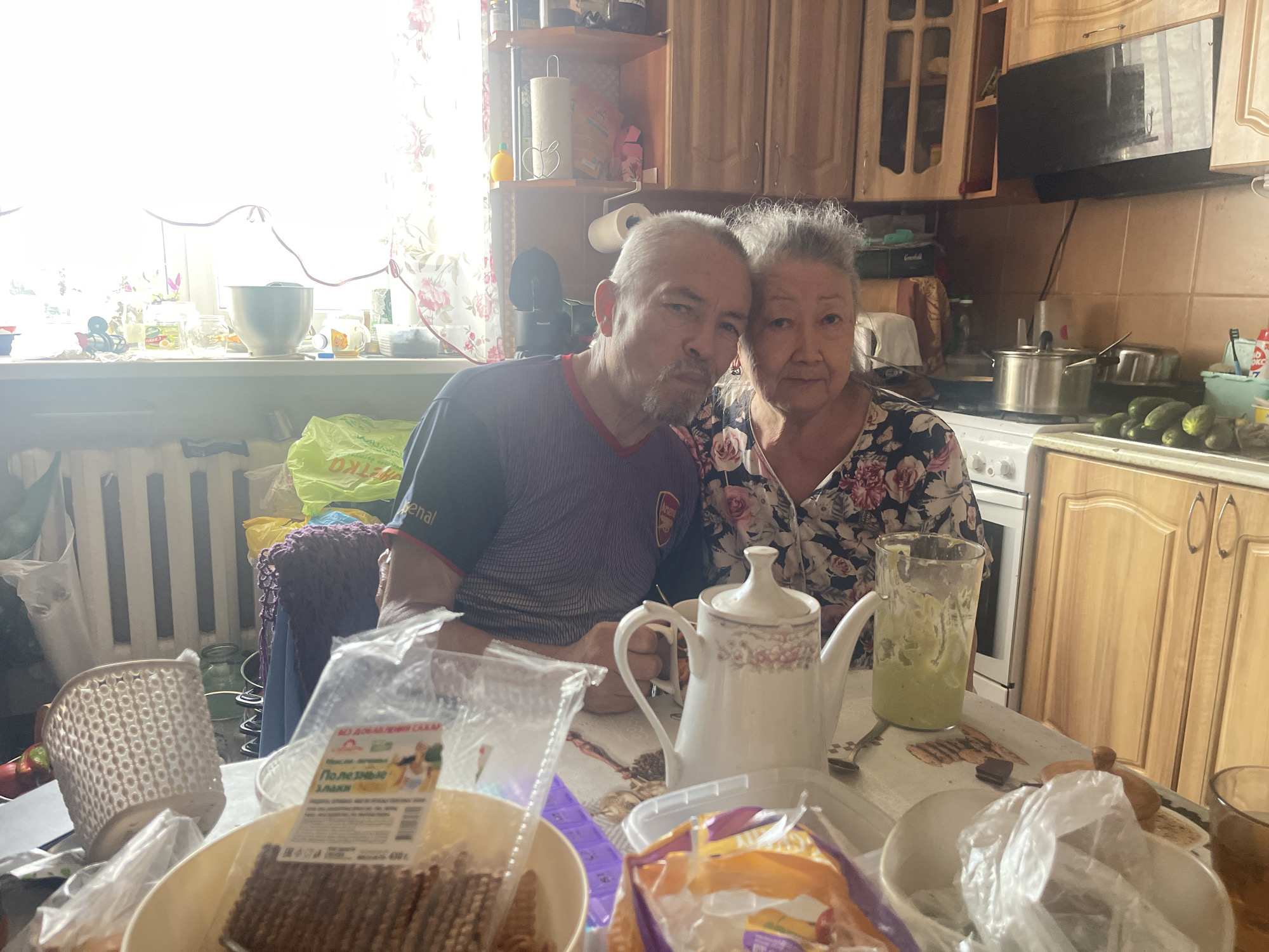 An indoor medium shot captures a man and a woman posing for a photograph in a kitchen. The man is on the left, facing forward with his head tilted slightly towards the woman, who is on the right. Both appear to be middle-aged or older. The man wears a purple and red t-shirt, while the woman is dressed in a floral-patterned blouse. They are seated at a table laden with various items, including a white teapot, a glass with green liquid, and packaged food items. In the background, kitchen cabinets and a stove are visible. The scene suggests a casual, domestic setting in Sibirskiy, Russia, possibly a home kitchen, as indicated by the presence of typical household items and the style of the cabinetry.