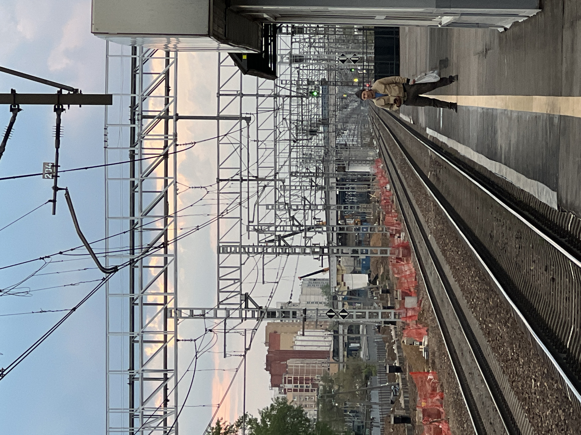 The image shows a train platform in Khimki, Russia. A man wearing a beige jacket and dark pants walks along the platform. He is carrying a white bag. The platform has a yellow safety line. The train tracks run parallel to the platform, and construction with orange fencing is visible near the tracks. Above the tracks, a complex network of overhead wires and metal structures spans the scene. In the background, buildings and trees are visible. The sky is overcast with soft pink and gray clouds.