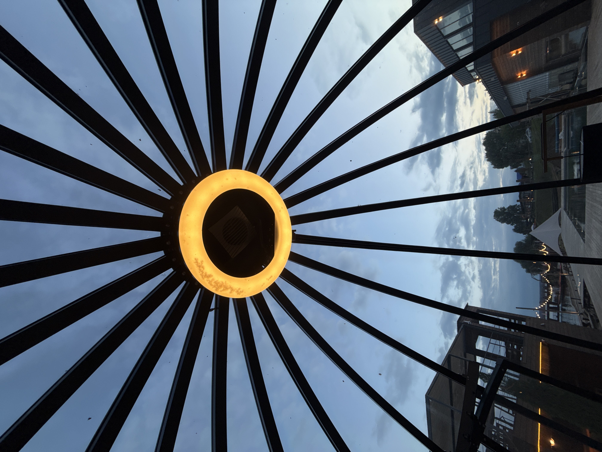 This media file captures a view from within a structure with a radial ceiling. The ceiling is composed of dark, thin beams radiating from a central point, creating a sunburst or spoke-like pattern. At the center of this pattern is a bright, circular light fixture, glowing with a warm yellow-orange hue. The light fixture appears to have a textured surface and is casting a warm illumination.
Through the transparent ceiling and surrounding windows, the sky is visible, displaying a cloudy, dusk or twilight scene. The clouds are soft and diffused, with hints of lighter tones where the sky is breaking through.
Beyond the structure, an outdoor scene is partially visible. This includes modern-looking buildings with illuminated windows, suggesting a contemporary setting. Trees and some landscape features can be discerned, along with what appears to be a body of water or a waterfront area. String lights are visible in the distance, adding to the ambiance of the evening. The overall mood is serene and somewhat atmospheric, characteristic of a peaceful evening or late afternoon in an outdoor or semi-outdoor venue.
The location context provided is Votkinsk, Russia. While specific landmarks are not clearly identifiable, the combination of modern architecture and a potentially recreational waterfront area suggests a developed public space. There is no visible text in the image. The activity appears to be a still observation of the environment, with no people or dynamic events depicted.