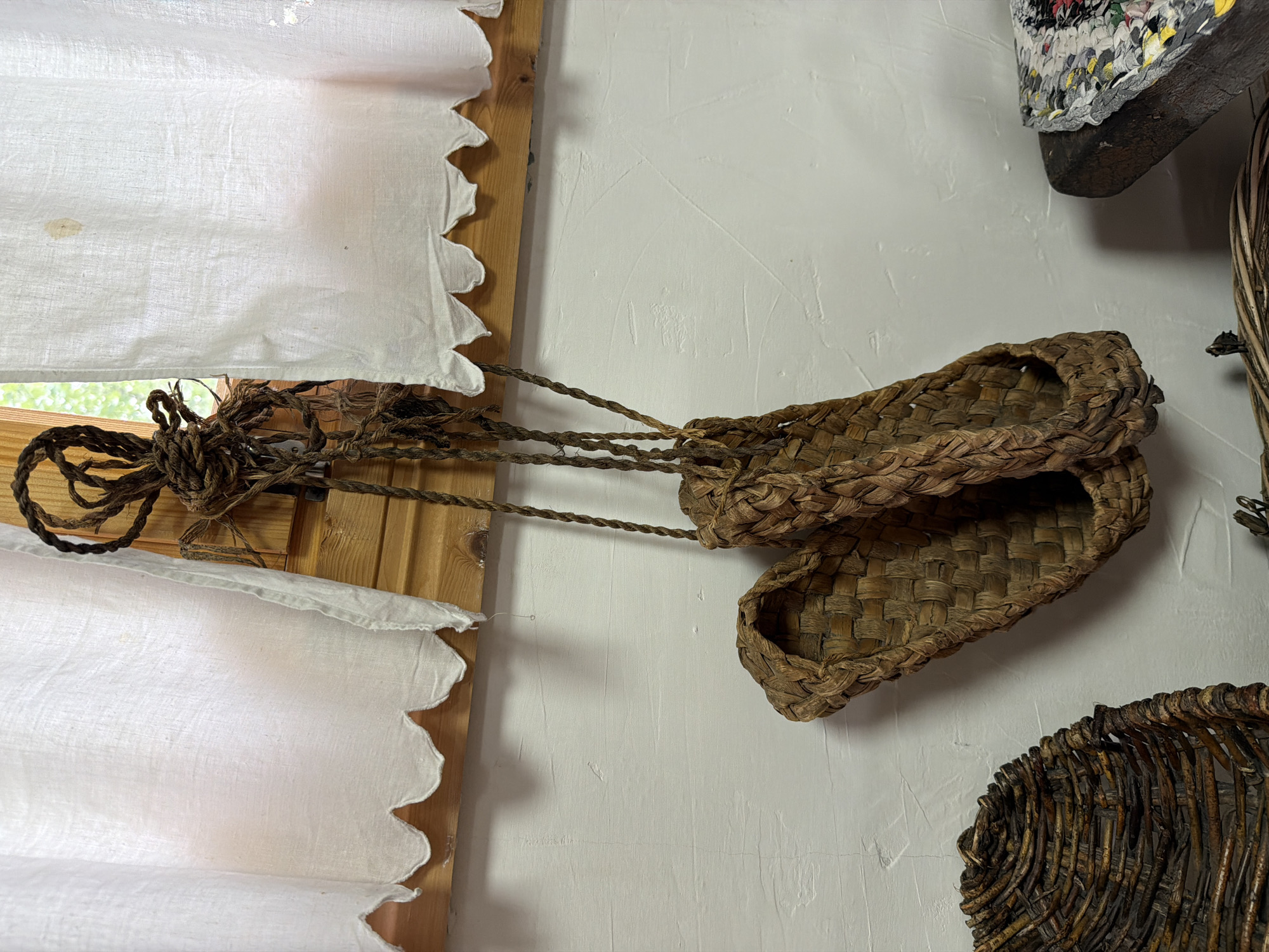 A pair of woven, natural fiber sandals hangs by their braided rope straps from a wooden support beam in front of a white, textured wall and a white linen curtain with a scalloped hem. The sandals are made of what appears to be straw or reeds, woven in a basket-like pattern, with a braided edge and a toe strap. They are positioned in the center of the frame, slightly angled. To the left, the edge of another woven basket is visible. To the right, a dark, rough-hewn wooden object supports a colorful woven mat or textile. The lighting suggests an indoor setting, possibly a rustic dwelling or museum exhibit, with natural light filtering in from a window behind the curtain. The overall impression is of handcrafted, traditional footwear displayed in a simple, unadorned environment.