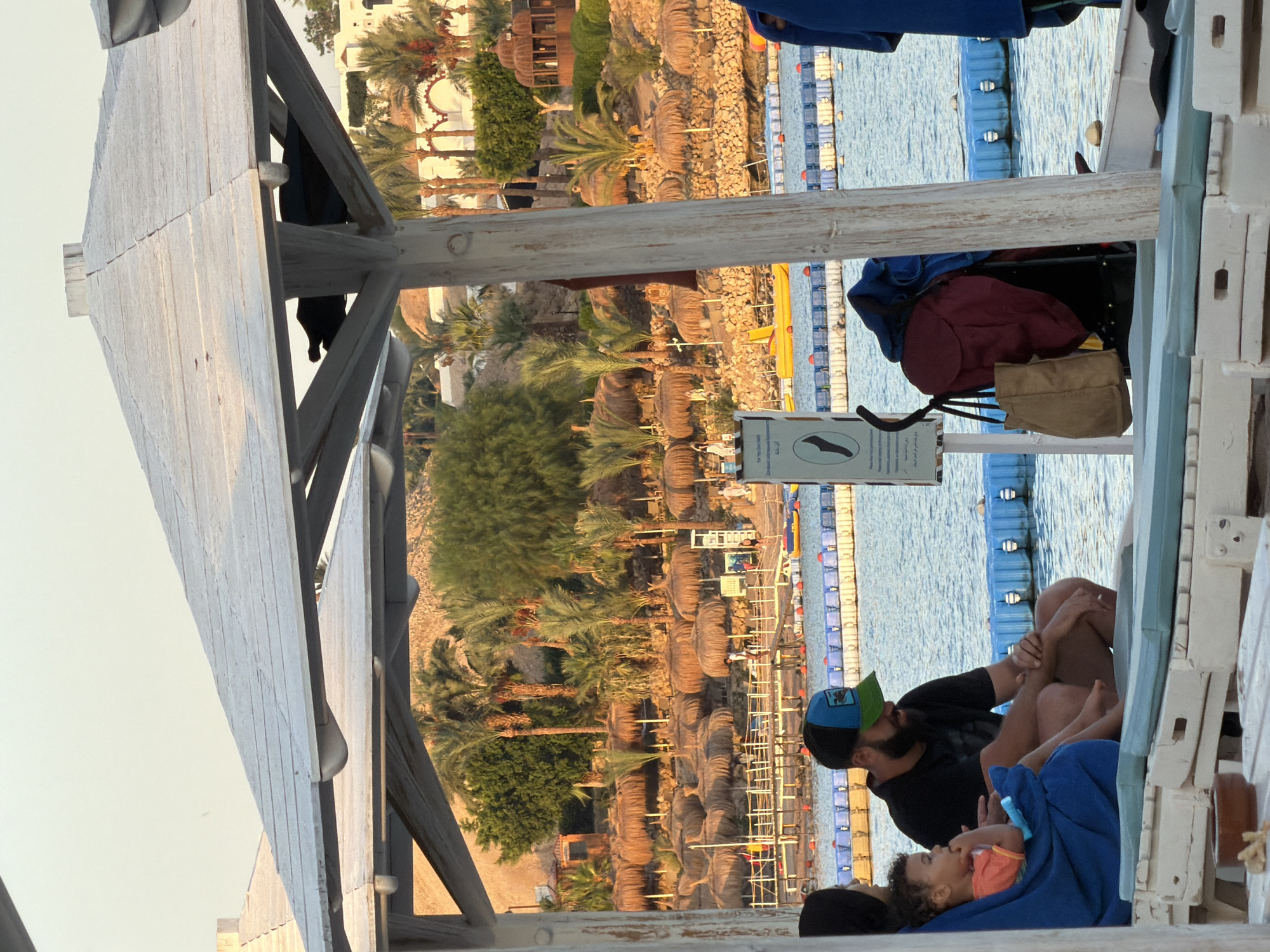 The image depicts a resort scene on a beach in Sharm El Sheikh, Egypt. In the foreground, a family is seated on lounge chairs under a white thatched-roof umbrella. A man wearing a black t-shirt and a green and blue baseball cap is seated next to a young child wrapped in a blue towel, who is wearing an orange shirt. Another person, mostly obscured by the umbrella, is seated to the left. A stroller with a burgundy bag and a tan tote bag is parked nearby.
The middle ground shows a body of water with a blue floating barrier. In the distance, a beach is lined with numerous thatched umbrellas and palm trees. Buildings with white facades and arched windows are visible on a hillside behind the beach. The sky is clear with a pale yellow hue, suggesting either sunrise or sunset. A sign with a foot icon and text in Arabic and English is mounted on a pole near the stroller. The overall atmosphere is relaxed and vacation-oriented.