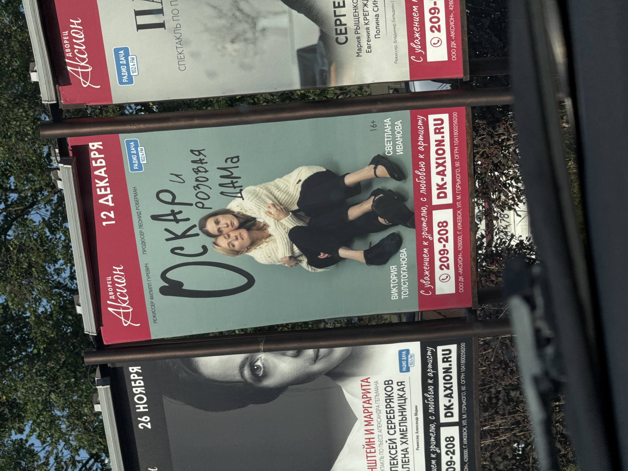 The image displays a series of three billboards in an outdoor setting, likely a street in Izhevsk, Russia, as indicated by the text "ИЖЕВСК" (Izhevsk) and street names on the billboards. The billboards are advertising theatrical productions.
The leftmost billboard, partially visible, features a black and white photograph of a woman's face and text that includes "26 НОЯБРЯ" (November 26) and "МАРГАРИТА" (Margarita). Below this, there is text mentioning "АЛЕКСЕЙ СЕРЕБРЯКОВ" (Alexei Serebryakov) and "АЛЕНА ХМЕЛЬНИЦКАЯ" (Alena Khmelnitskaya), along with "РЕЖИССЕР: Александр Морин" (Director: Alexander Morin). The bottom of this billboard shows contact information including a phone number and "DK-AXION.RU".
The central billboard is the most prominent. It has a red header with "ДВОРЕЦ АКЦИОН" (Palace of Action). The main part of the billboard is light green and features the title "ОСКАР и РОЗОВАЯ ДАМА" (Oscar and the Pink Lady) in large, stylized font. Below this, there are images of two women embracing. Text identifies "ВИКТОРИЯ ТОЛСТОГАНОВА" (Victoria Tolstoganova) and "СВЕТЛАНА ИВАНОВА" (Svetlana Ivanova). A "16+" rating is also present. This billboard also advertises "12 ДЕКАБРЯ" (December 12) and includes "РАДИО ДАЧА 102.4 FM" (Radio Dacha 102.4 FM). At the bottom, it reads "С уважением к зрителю, с любовью к артисту" (With respect to the audience, with love to the artist), followed by a phone number and "DK-AXION.RU", with additional address details including "г. ИЖЕВСК, ул. М. ГОРЬКОГО, 92" (Izhevsk, M. Gorky Street, 92).
The rightmost billboard, also partially visible, has a red header with "ДВОРЕЦ АКЦИОН" and text indicating "СПЕКТАКЛЬ ПО П..." (Spectacle by...). It features the name "СЕРГЕ..." (Serge...). Below this, there are names such as "Мария Рыщенкова" (Maria Ryshchenkova), "Евгения КРЕГЖД..." (Evgenia Krezhd...), and "Полина СИН..." (Polina Sin...). It also mentions "Режиссер: Владимир Бельдягин" (Director: Vladimir Beldyagin). The bottom of this billboard contains contact information including a phone number and "ООО ДК «АКСИОН»..." (LLC DK "Axion"...).
The billboards are mounted on poles and are positioned along a street, with trees visible in the background. Part of a car's windshield and dashboard are visible in the foreground, suggesting the photo was taken from inside a vehicle. The sky is clear and blue.