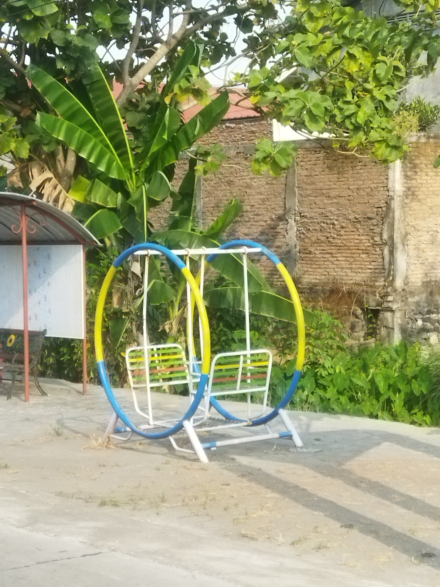 An outdoor scene in Juwiring, Indonesia, captures a colorful double swing set with blue and yellow circular frames, each supporting a white swing seat with green and yellow stripes. The swing set is situated on a paved area with scattered patches of dry grass and dirt. Behind the swing set, a textured brick wall forms the backdrop, with lush green foliage and large-leafed plants growing in front of it. To the left of the swing set, a red metal bench with a decorative design is visible under a simple, open-sided shelter with a corrugated metal roof. The overall environment suggests a public space or a backyard area in a tropical or subtropical climate, likely during daylight hours given the lighting and shadows.