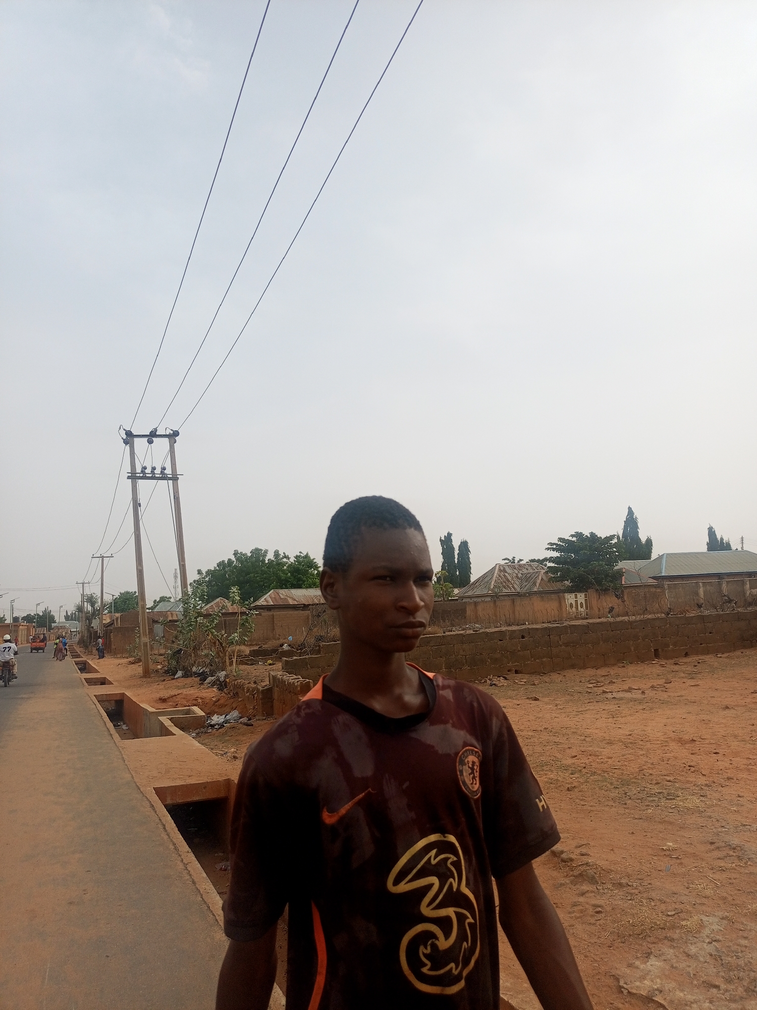 A young man stands in the foreground of a semi-urban or rural street scene in Tempure, Nigeria, looking slightly to his right with a neutral expression. He wears a dark, possibly maroon or brown, Chelsea FC football jersey featuring a large golden "3" logo on the front, a white Nike swoosh, and the club badge. Part of "HYUNDAI" is visible on his right sleeve.
The sky is bright, hazy, and overcast, suggesting daylight hours but without harsh shadows, possibly indicating a dry or dusty season. Overhead, multiple power lines crisscross the frame, connecting to a series of wooden utility poles that stretch into the mid-ground.
To the young man's left, a paved road extends into the distance, lined by open concrete drainage channels. Further down the road, several people are visible, including a person riding a motorcycle (okada) with a passenger whose white shirt has the number "77" printed on the back.
On the right side of the image, the ground is reddish-brown dirt with scattered dry vegetation and some debris. Beyond this, low concrete block walls and various modest houses with corrugated tin roofs and exposed brick walls are visible. Tall, slender trees dot the distant landscape, characteristic of the local environment.