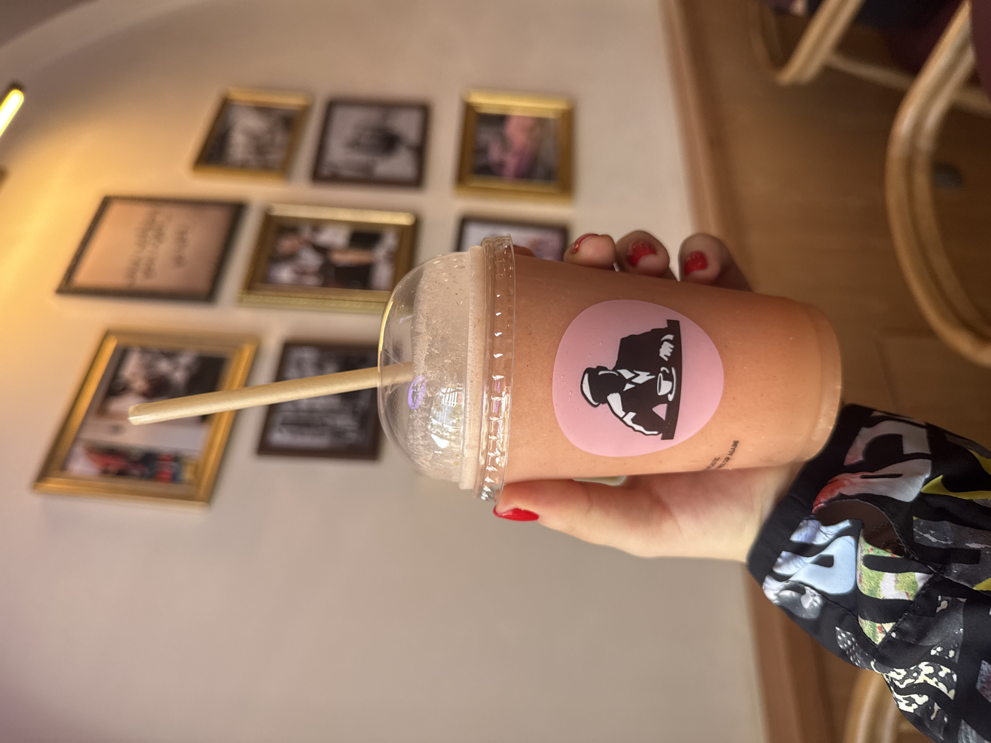 A person's hand, adorned with red nail polish, holds a light orange-colored beverage in a clear plastic cup with a dome lid and a straw. A circular light pink sticker on the cup displays a black and white logo featuring a stylized figure, possibly a person in a hat, sitting at a table with a drink. Below the logo, partial text "ATIC" and "UP AGAIN" is visible. The person is wearing a multi-colored, patterned jacket sleeve.
The setting is an indoor space, likely a cafe or restaurant in Mexico City, Mexico. The background features a light-colored wall decorated with an array of framed pictures, some with golden frames, others darker. These frames appear to hold various photographs, some in black and white, and at least one containing blurry text. A bright, linear light fixture illuminates the upper left corner of the scene. A blurred wooden object, possibly a piece of furniture, is visible on the right side of the frame. The overall impression is that of a casual moment, enjoying a drink in a decorated establishment.