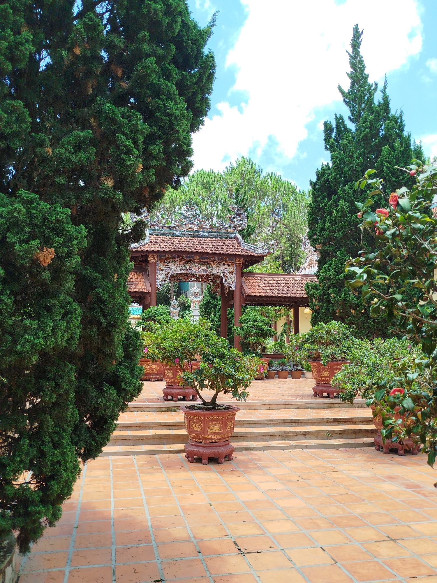 The image shows a courtyard in the city of Da Lat, Vietnam. The courtyard is paved with square orange tiles, and is lined with trees and potted plants. In the background, there is a traditional Vietnamese pavilion with a red tile roof. The sky is blue with scattered clouds.