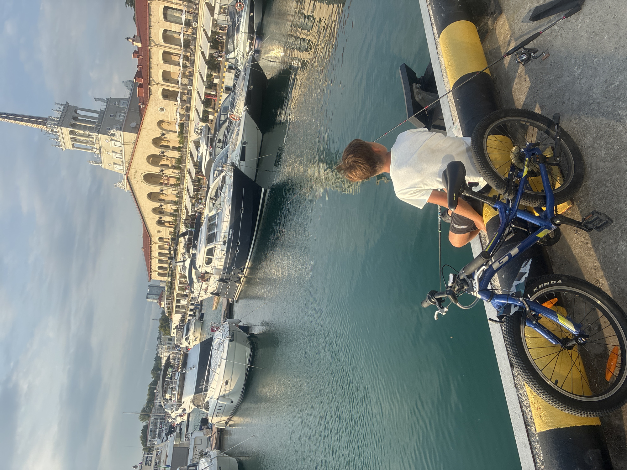 In Sochi, Russia, a young boy is seen fishing from the dockside, with his bicycle resting beside him. The scene unfolds at the Sochi Sea Port, characterized by its iconic Stalinist architecture, featuring a grand building with arches and a prominent tower. The water in the port is a clear, turquoise hue, reflecting the sunlight. Several yachts and boats are moored in the harbor, adding to the maritime atmosphere. The boy, wearing a white t-shirt and shorts, is engrossed in his fishing activity, holding a fishing rod. The image captures a tranquil moment in a bustling port city.