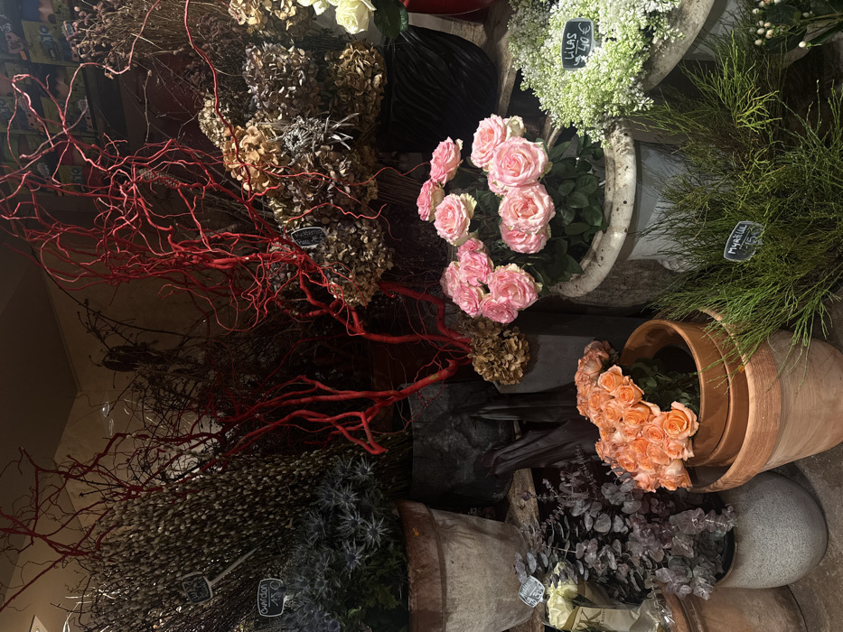 A vibrant display of flowers and foliage in varying pots and containers. Pink roses, peach-colored roses, and white blooms are visible. Dried hydrangeas, red branches, and feathery green plants add texture. The containers range from terracotta pots to aged stone vessels. Small signs with handwritten prices are attached to some arrangements. The overall composition creates a rustic and charming floral shop scene.