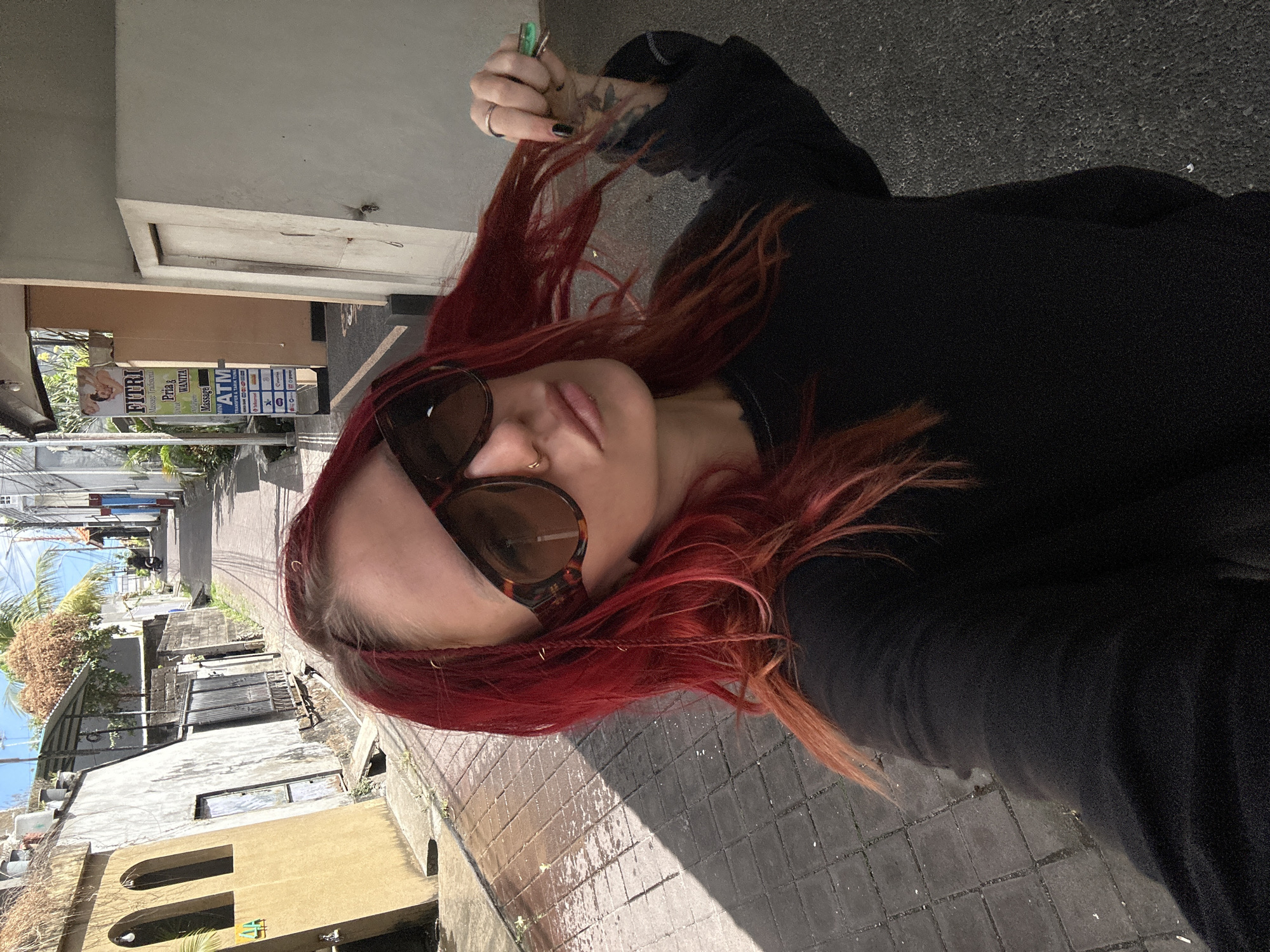 A woman with bright red hair and large, patterned sunglasses is standing on a narrow, paved street. She is wearing a black long-sleeved shirt and is holding a strand of her hair. The street is lined with buildings, some of which have arched windows. A sign for "FITRI Massage Tradisional" is visible on one of the buildings, as well as an ATM. The street appears to be in a slightly downhill direction. The sky is clear and blue.