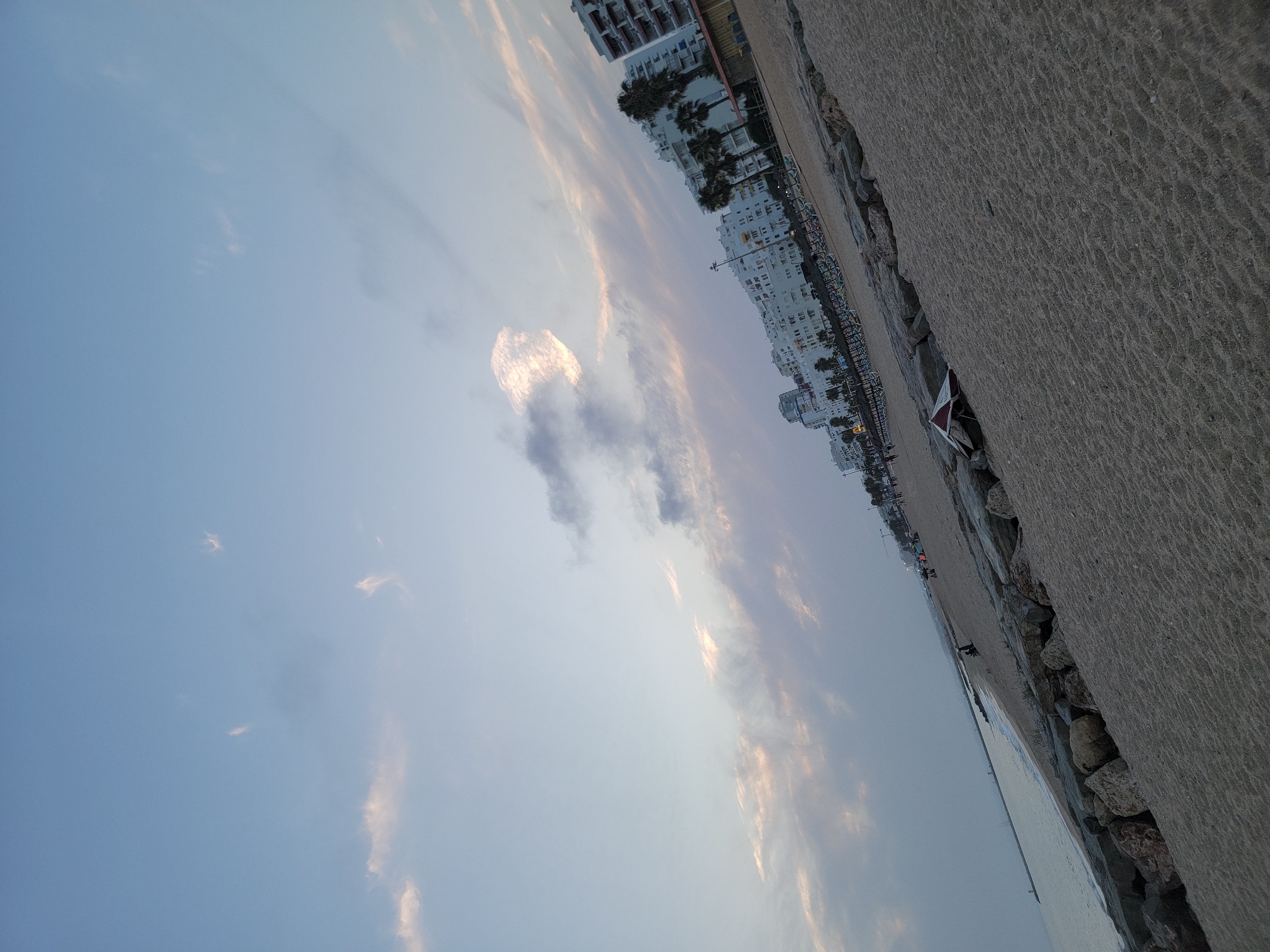 The image depicts a slightly tilted view of Quarteira Beach in Quarteira, Portugal, during what appears to be the late afternoon or early evening. The sky is a soft blue with wispy clouds, some tinged with pink and orange hues, suggesting the sun is low on the horizon.
The foreground is dominated by coarse, textured sand. A line of large, rough-hewn rocks forms a barrier along the edge of the sand, separating it from the sea. Beyond the rocks, the calm, light-colored water of the Atlantic Ocean meets the shore.
In the mid-ground, a promenade or boardwalk is visible, lined with rows of empty beach chairs and umbrellas, indicating the beach is likely winding down for the day. Behind the chairs, a series of white, multi-story buildings, characteristic of coastal resorts, are visible. These buildings have balconies and windows, and some are topped with red-tiled roofs. Palm trees are interspersed between the buildings and along the promenade.
Further down the beach, a few individuals can be seen as small figures, either walking or sitting. A distant pier or breakwater extends into the sea on the left side of the image, marked by a white structure. The overall impression is one of a serene, populated beach at the close of day.