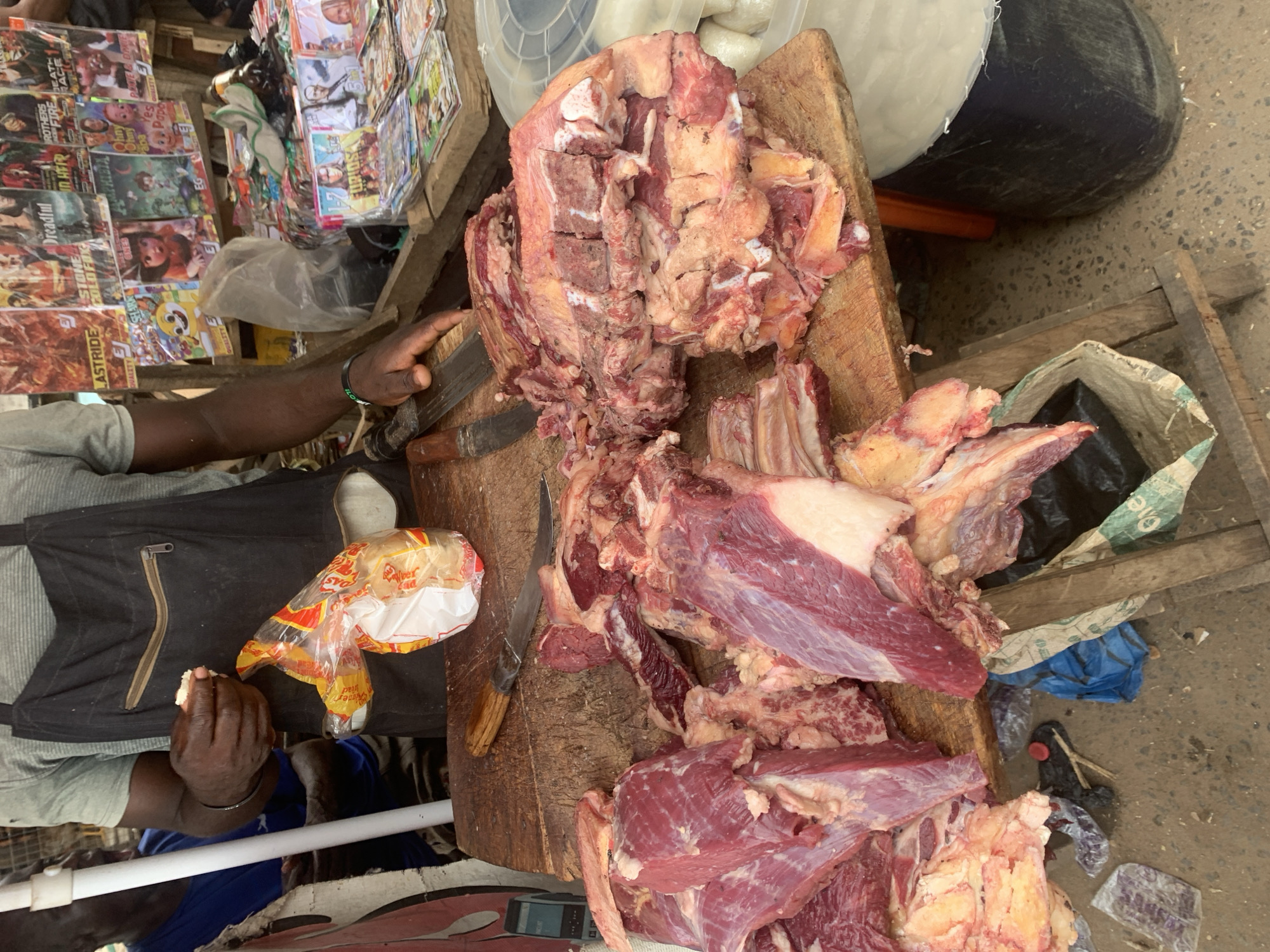 This image captures a vibrant market scene in Shagamu, Nigeria, centered around a meat vendor's stall. A person with dark skin, wearing a grey t-shirt and a black apron, is visible on the left. Their left hand holds a clear plastic bag containing bread, while their right hand is near a large knife with a wooden handle on a wooden cutting board.
The main subject is a substantial display of raw meat, likely beef, piled on the robust wooden cutting board. The meat shows various fresh cuts, including muscle, bone, and fat, ready for sale or further processing.
In the background, on the left, are shelves or a stall displaying numerous printed materials such as magazines, comic books, or DVDs. Visible titles and phrases include "LASTRIDE," "QUALITY," "STONE COLD FIX," "Dreadful," "BROTHERS IN THEIR FIRE DEATH RACE," "AMPHIBIA," "ONES," "FURIOSA," and "Ohny and Yes Papa."
Behind the meat stall, to the right, are a white plastic container and a dark-colored bin, with a woven sack lying on the ground below. The ground is a rough concrete or dirt surface. The scene is set outdoors during daylight hours, reflecting a busy, functional market environment. A black wristband on the vendor's right arm has "iFLow" visible. The bread bag shows partial text like "Silver Warrier."