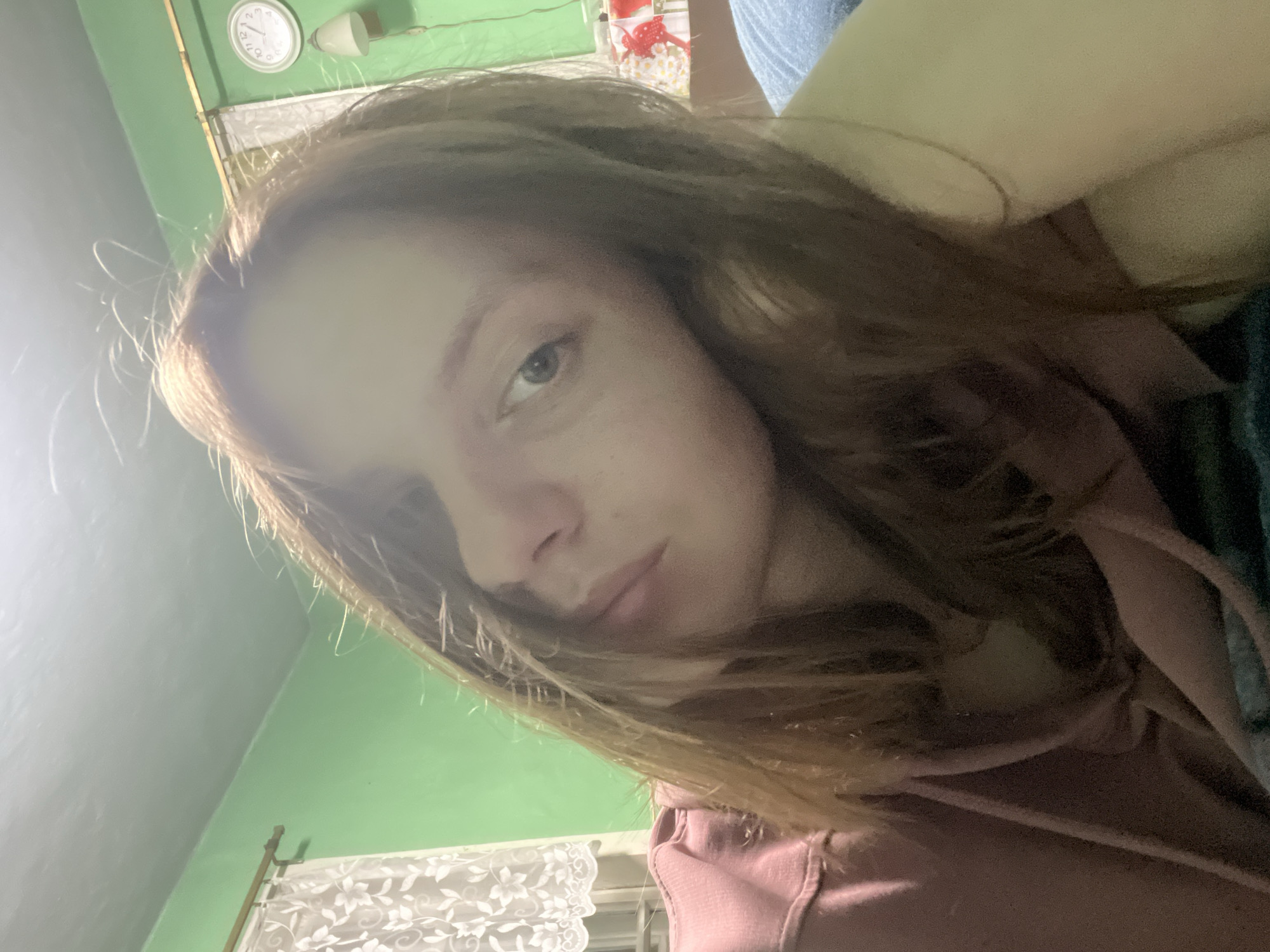 This is a close-up, angled selfie of a young woman with long, light brown hair. She is wearing a pink hooded sweatshirt. Her face is partially obscured by her hair, but her left eye and part of her nose and mouth are visible. She has a neutral expression.
The background is an indoor scene. To the upper left, a white clock with black Roman numerals is visible on a light green wall. To the right of the clock is a white light fixture. Below the clock and to the left, a white lace curtain is visible. The overall lighting suggests it might be indoors with artificial light. The location is specified as Nizhny Tagil, Russia. No specific activity or event is apparent beyond the act of taking a selfie.