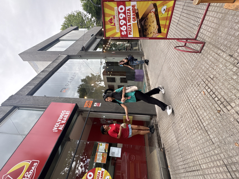 The image captures an urban street scene on an overcast day in Villa Luro, Buenos Aires, Argentina, specifically at Cnel. Ramón L. Falcón 4899. The foreground features a light grey tiled sidewalk.
On the left, a building with a prominent red facade displays a partial sign reading "POLLO ASADAS" and "¡VOLVIMOS AL BAILE!" above a glass entrance. Inside the doorway, a female with dark hair, wearing a red t-shirt and a light blue denim skirt, stands facing outwards, holding a small white bag in her left hand. A red vehicle is reflected in the glass entrance.
Adjacent to the red-fronted building, to the right, stands a structure with a dark grey, textured facade and large glass windows that reflect the street and sky. The text "RAMAT IMPORTS" is visible on the glass. A string of decorative lights hangs beneath a canopy extending from this building.
Three females are present. The first, previously described, is inside the red building's doorway. The second female, with long dark hair, wearing a teal short-sleeved top, black wide-leg pants, and white Adidas sneakers, walks right to left across the mid-ground. She carries a light-colored tote bag over her left shoulder and holds small items in her left hand. Her gaze is directed downwards. The third female, visible further down the sidewalk on the right, also with dark hair, wears a dark top, ripped blue jeans, and black boots. She carries a dark backpack and is actively engaged in a phone call, holding a dark phone to her right ear while walking left to right.
On the far right, a yellow and red sandwich board sign stands on the sidewalk, advertising "$6990 PIZZA MUZZA GRANDE" and displaying an image of a large pizza. A red metal bicycle rack is partially visible in front of this sign. Trees with green foliage are visible in the background, rising above the buildings.