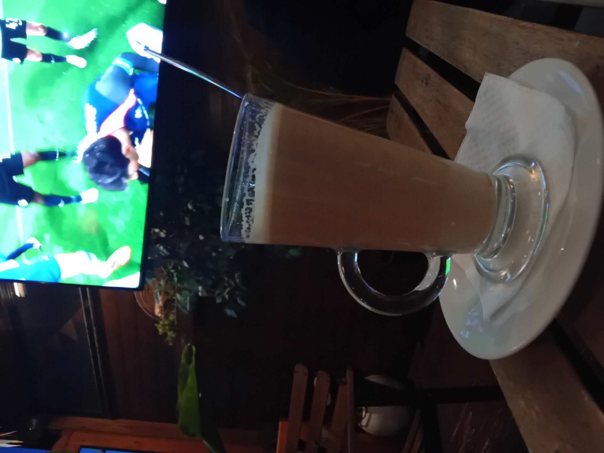 An indoor scene captures a tall glass of a light-colored beverage, likely a latte or coffee with milk, resting on a white saucer with a small white napkin underneath. The glass has a handle and is positioned on a wooden slatted table or surface.
In the background, a large screen displays what appears to be a soccer match. Figures in athletic attire, primarily blue and black, are visible on the bright green field of the screen. One player in a blue jersey is seen bending over. The screen is illuminated, suggesting the ambient lighting is dim, possibly indicating evening or nighttime.
Other background elements are dark and out of focus, with hints of wooden furniture and some dark greenery or plant decor. The overall setting appears to be a casual venue like a pub, cafe, or restaurant, where patrons can watch sports. No discernible text is visible in the image.