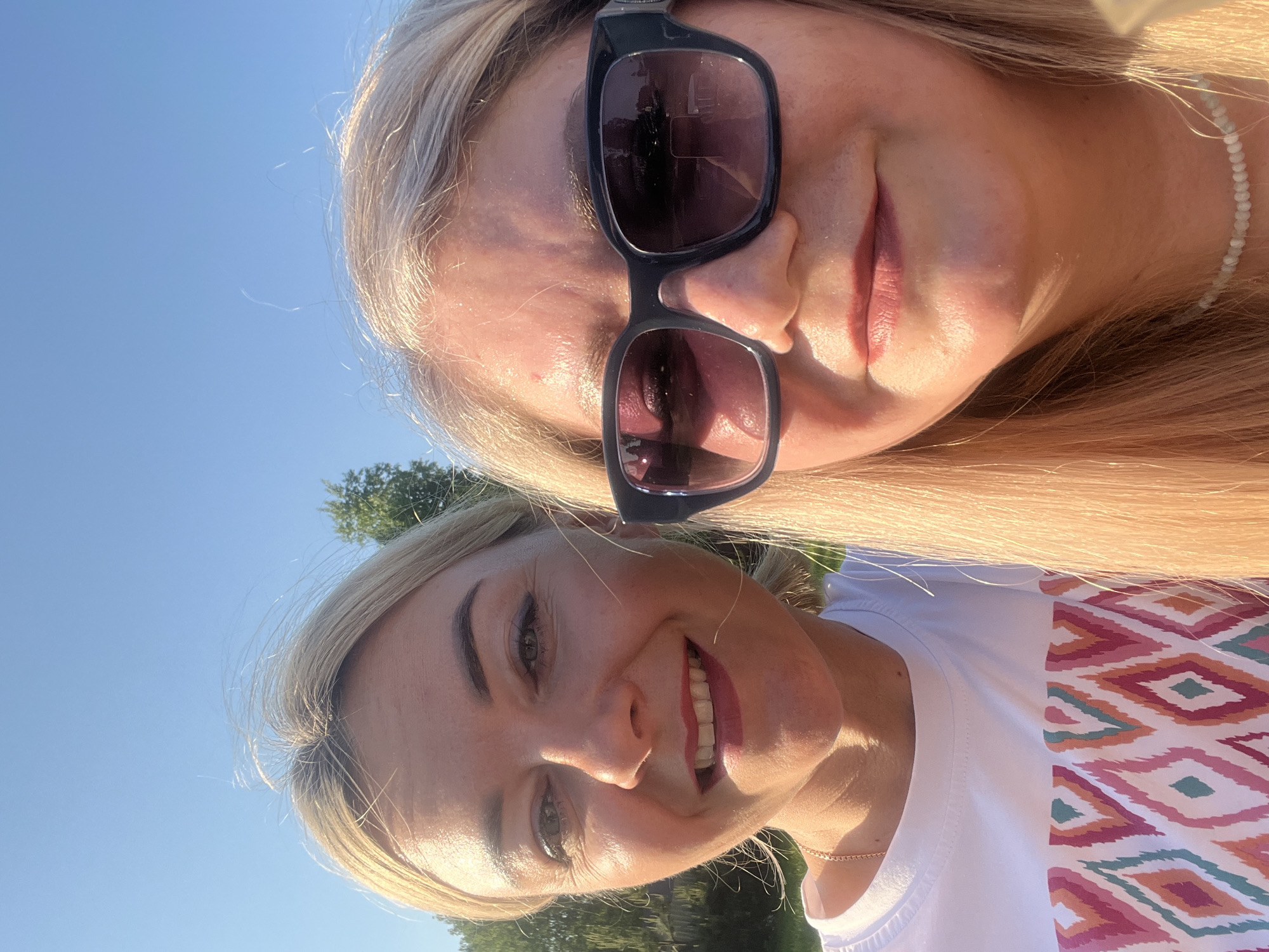 Two women, both fair-skinned with blonde hair, are posing close together outdoors. The woman at the top wears dark sunglasses and a light-colored beaded necklace. The woman at the bottom is smiling, wearing a white t-shirt with a geometric pattern. They are likely in Новая Чернушка, Russia. The background features a clear blue sky and some greenery, suggesting a park or outdoor setting. The photograph appears to be taken with a mobile phone.